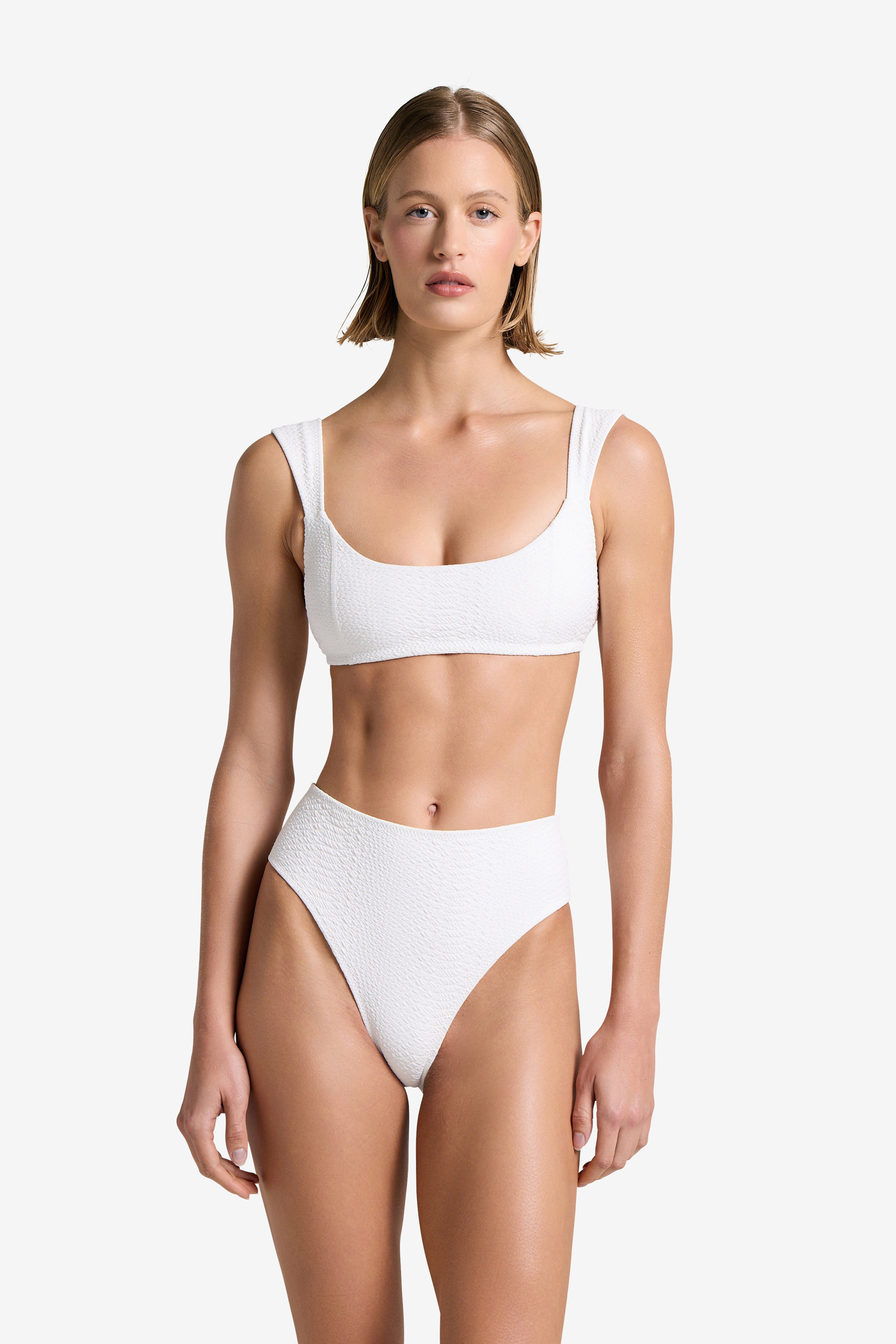 A person models the Dawson Top by Gigi C Bikinis, a white textured faux snake bikini with a scoop neckline and high-waisted bottoms, against a plain background. The bikini includes removable bust pads. Their expression is neutral, with straight shoulder-length hair.