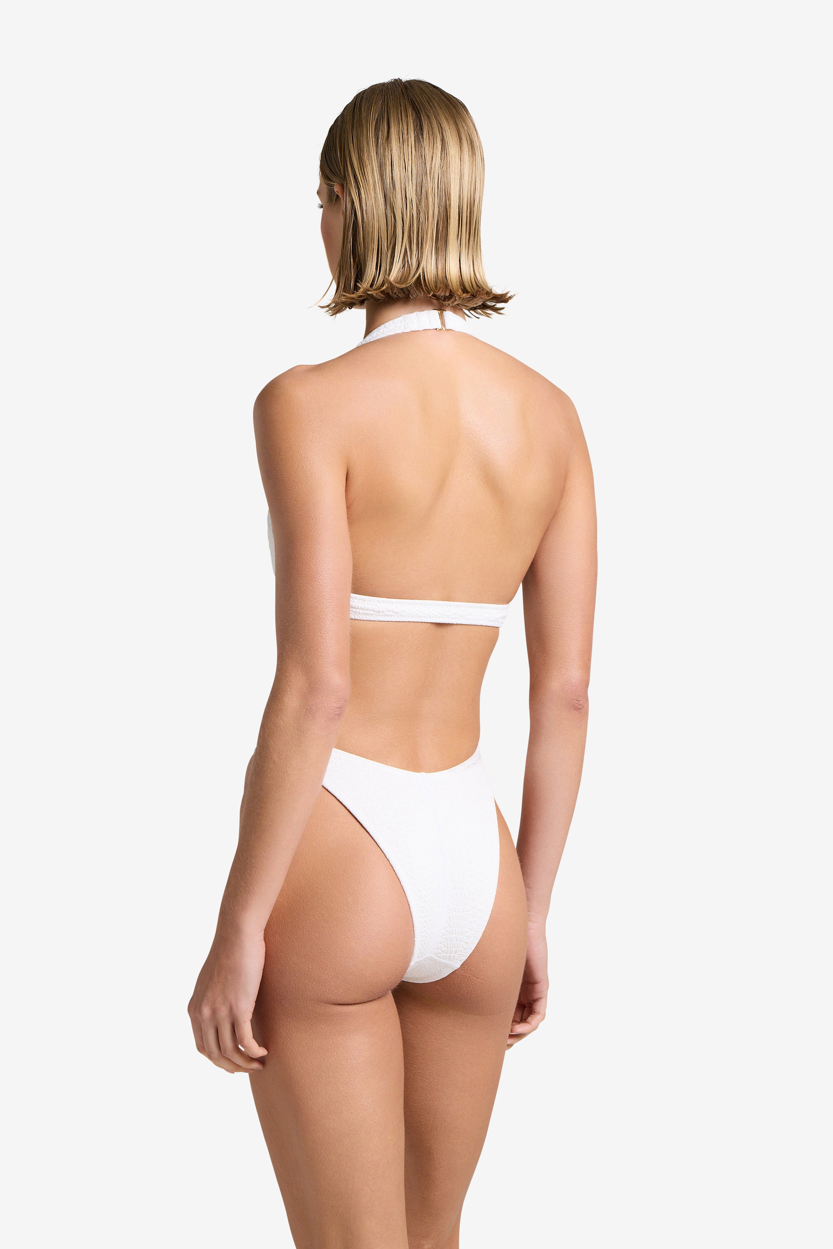 In a white setting, an individual with short blond hair models Gigi C Bikinis Megan One Piece. This stylish white halter-neck swimsuit, accented with gold hardware and featuring a high-cut design, elegantly showcases their back.