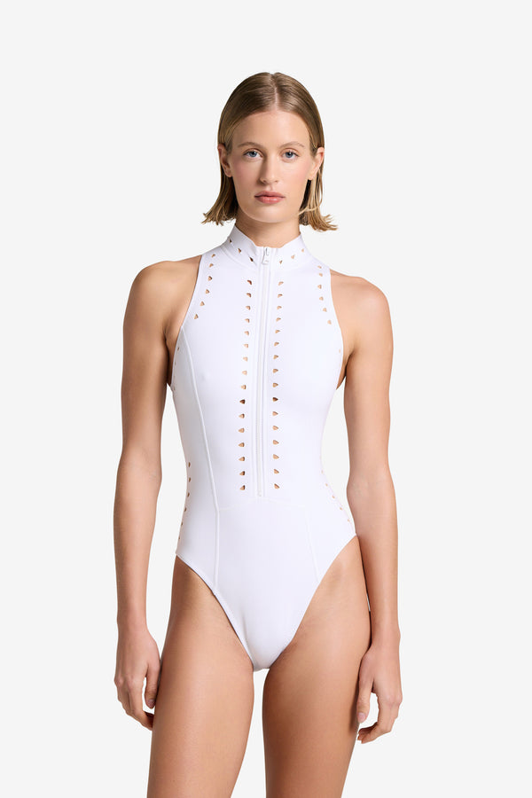 A person with short hair stands against a plain white background wearing the Mathilda One Piece swimsuit from Gigi C Bikinis, featuring a front zip closure and vertical cutout patterns on the front and sides, exuding a sleek scuba-inspired style.