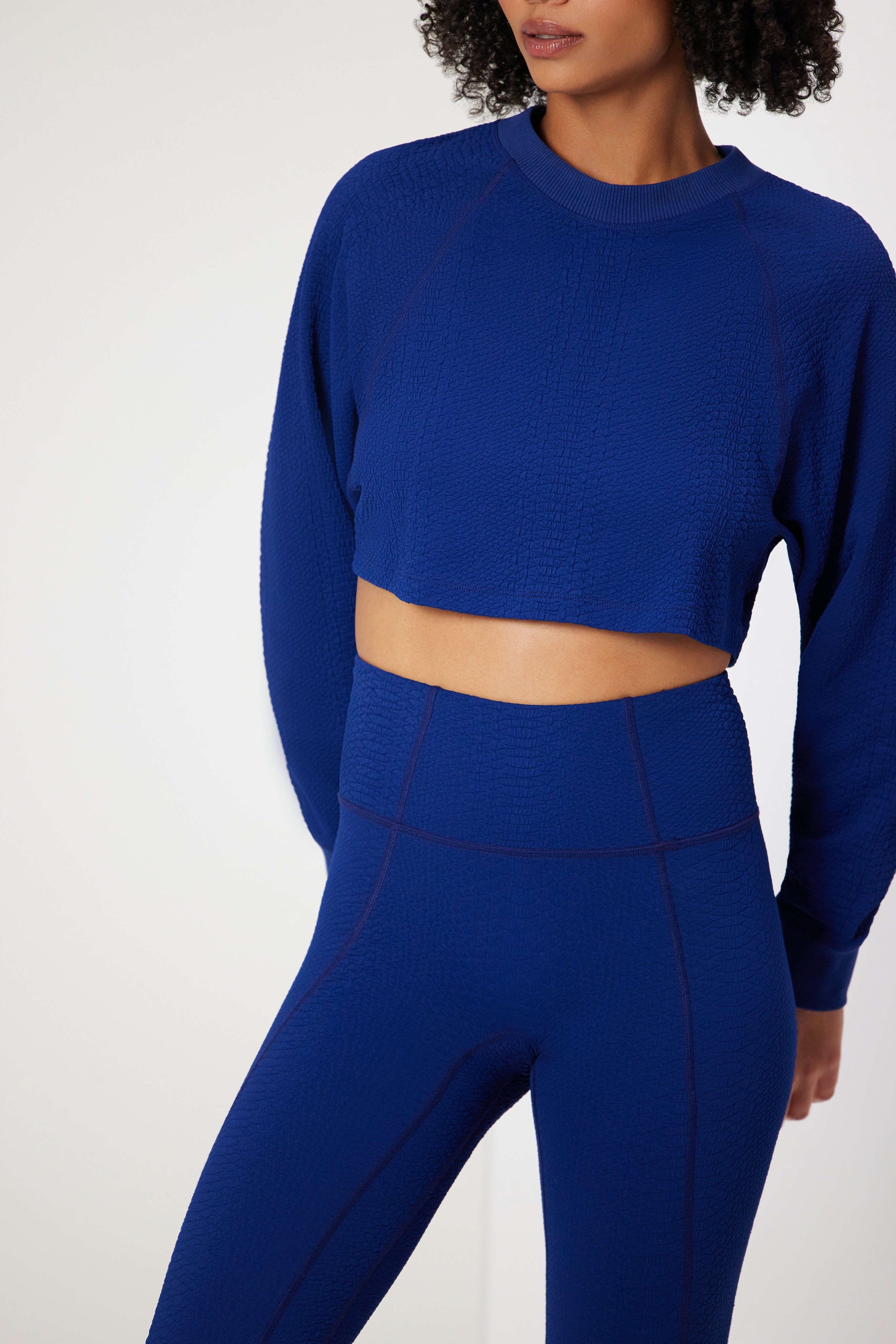 Wearing a textured blue crop top and high-waisted Ava Legging made from compression waffle jacquard by Gigi C Sport, a person stylishly showcases the form-fitting activewear. With curly hair framing their face, the photo beautifully highlights their upper body and outfit.