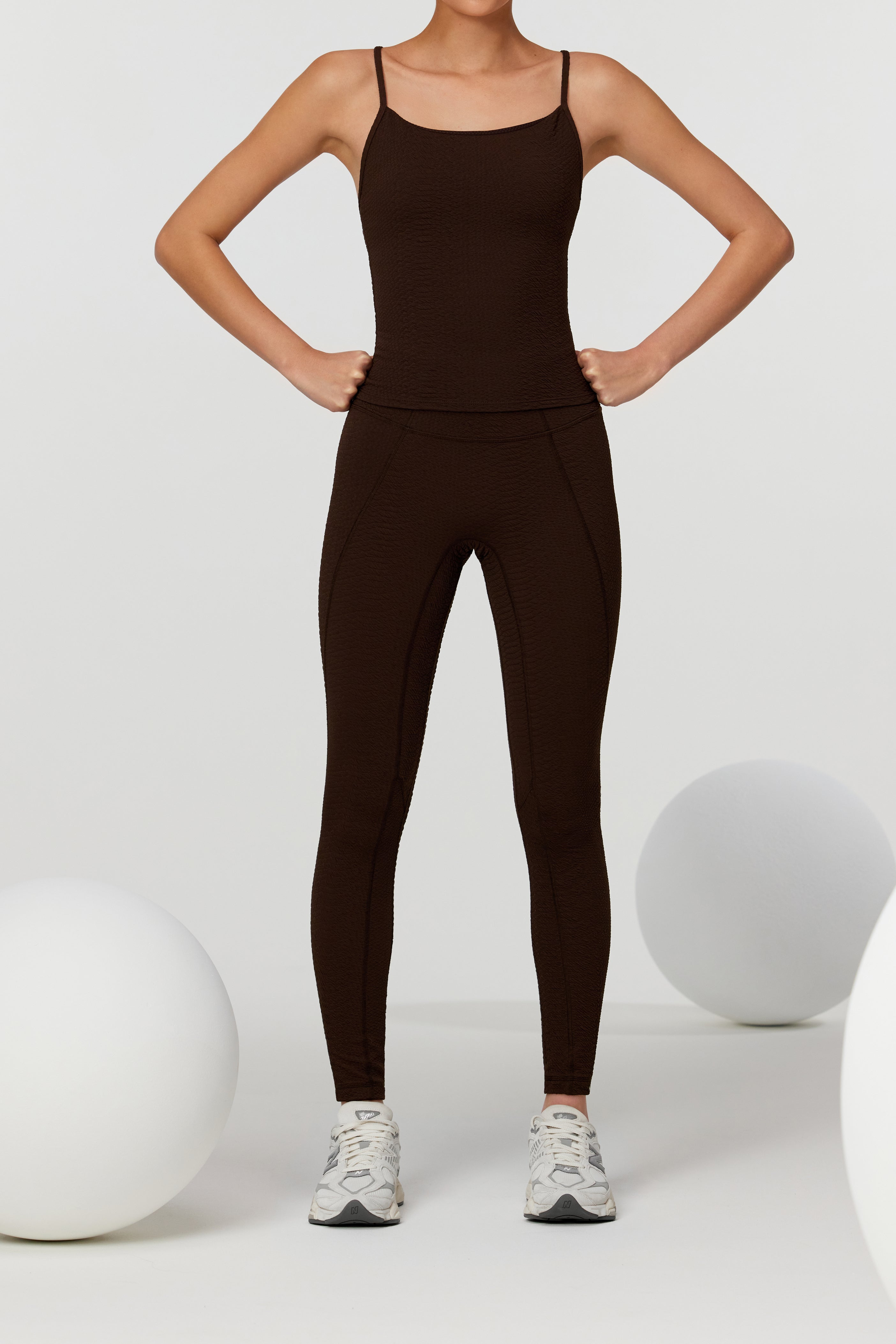 A person in a dark brown Ava Legging by Gigi C Sport and white sneakers stands confidently, hands on hips. The minimalist vibe is enhanced by a light gray background with two large white spheres beside them.