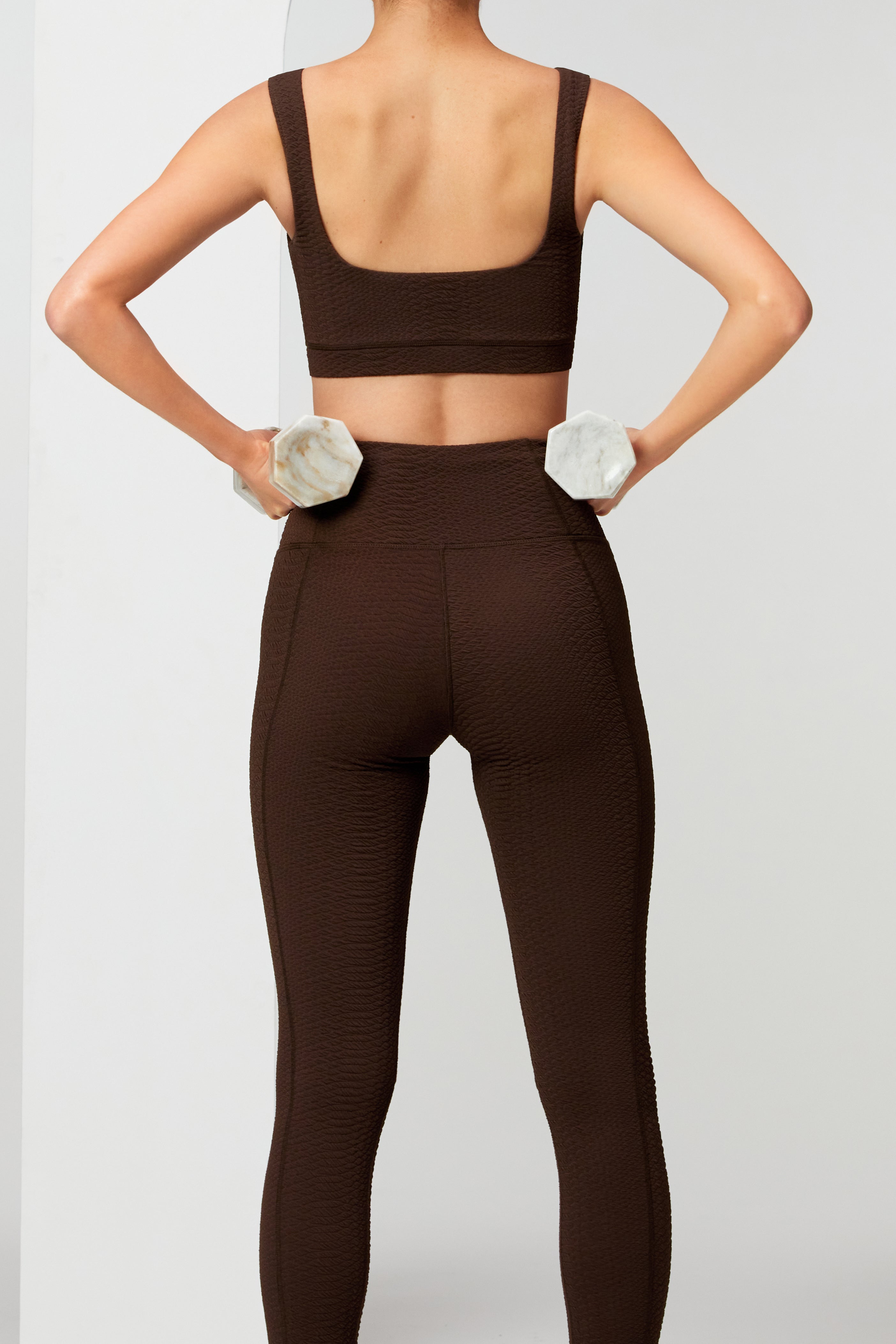 A person in a faux snakeskin sports bra and Ava Legging by Gigi C Sport stands facing away, holding marble weights with hands on hips. The workout attires fit is highlighted against a plain white wall.