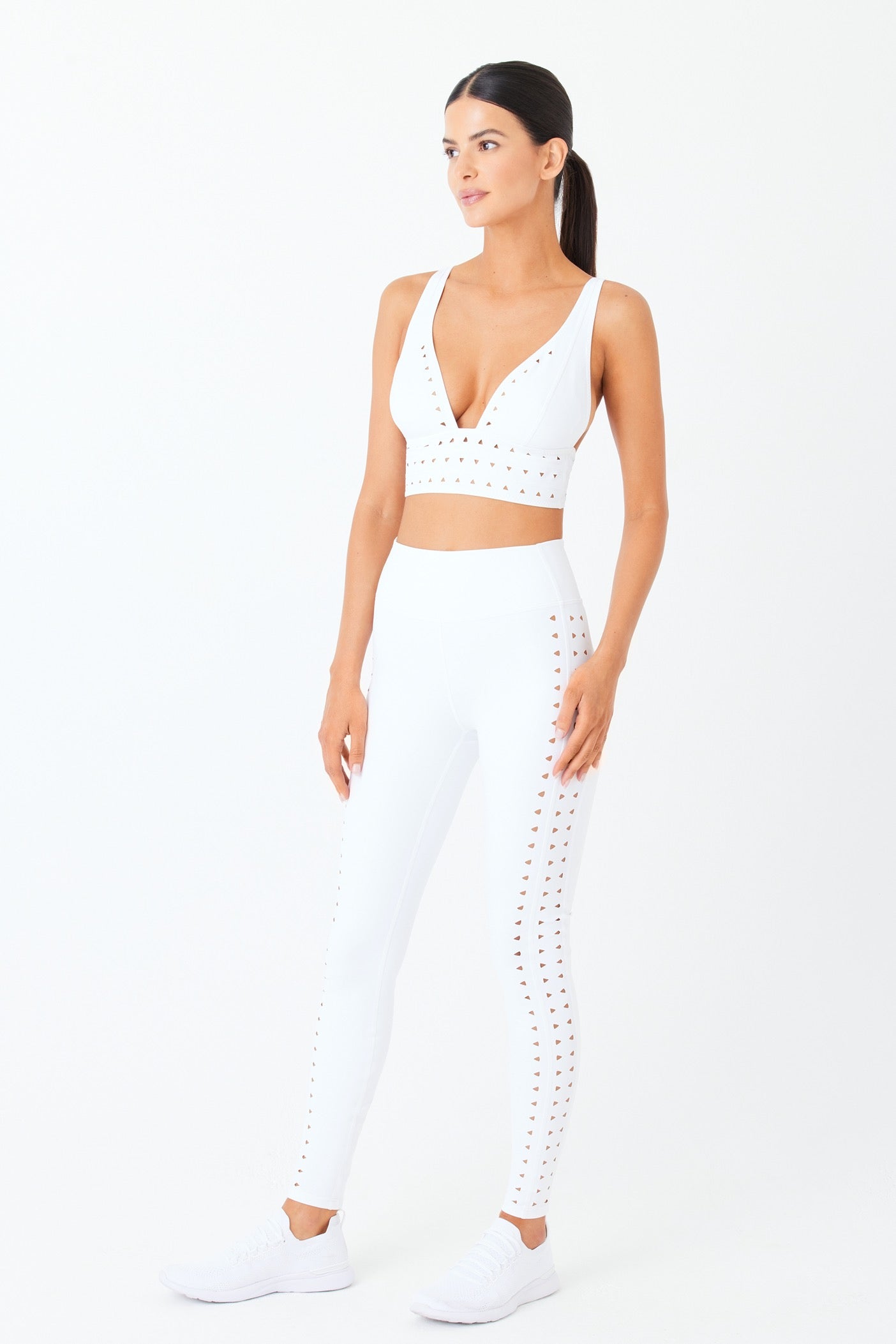 Wearing a Gigi C Sport Eden Bra and leggings, featuring precise cut-out details from lasercut technology, a person showcases the sleek white sports ensemble with breathable fabric sneakers, their long dark hair styled back against a plain white background.