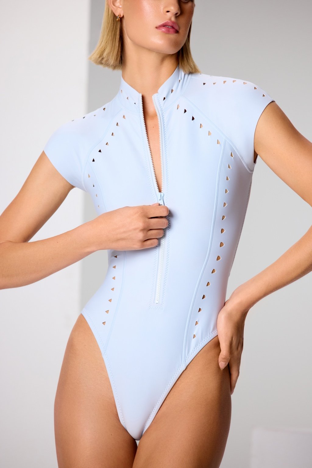 Clad in a light blue, short-sleeved Lydia Surfsuit by Gigi C Bikinis, with a front zipper and eyelet detailing in scuba fabric, they stand confidently with one hand on their hip and the other near the zipper in a bright, minimalist setting.