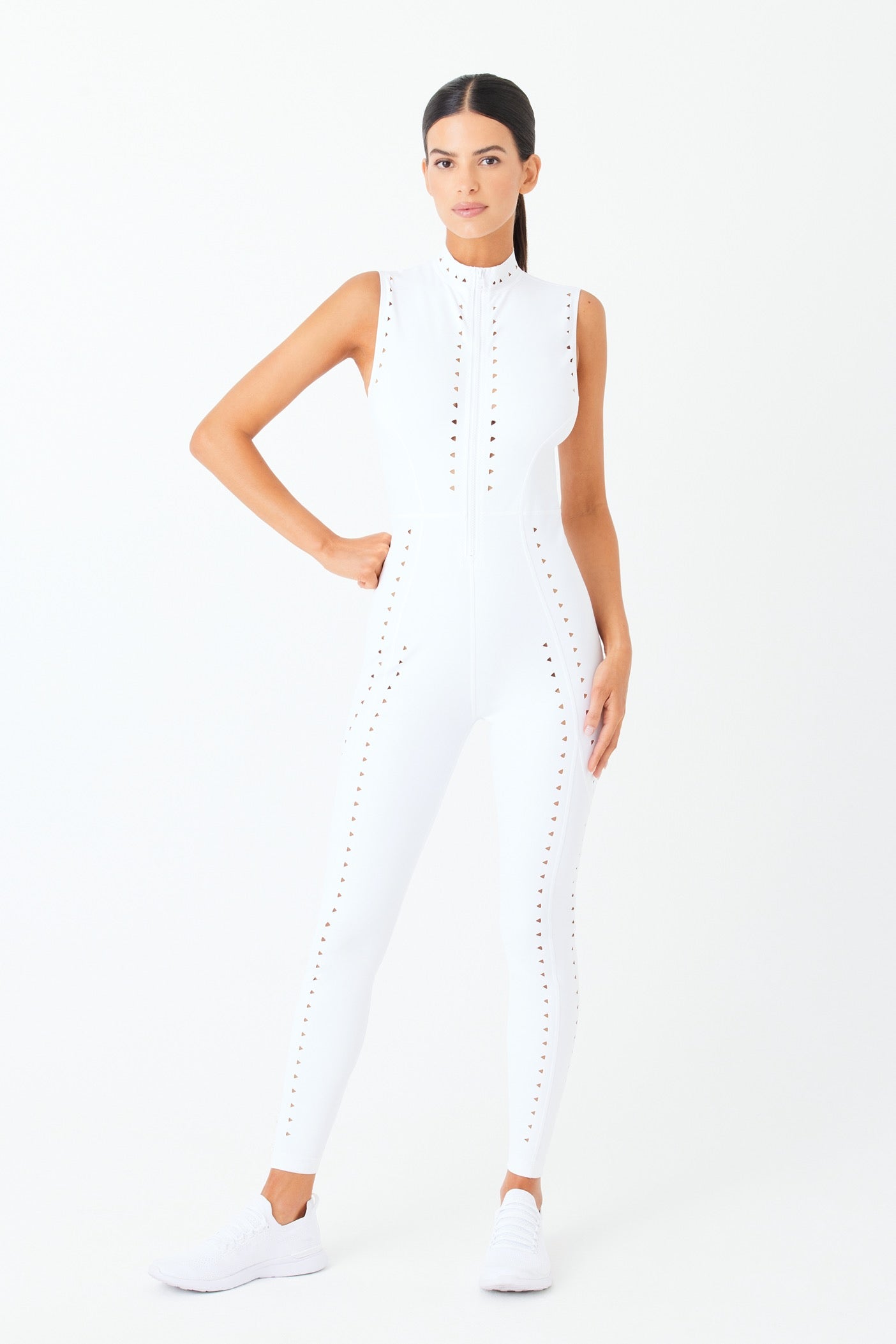 A person poses in a fitted, sleeveless Gigi C Sports Sarah Jumpsuit with laser cutout designs on the front and sides. They have long dark hair, wear white sneakers, and stand against a plain white background, emphasizing the chic look.