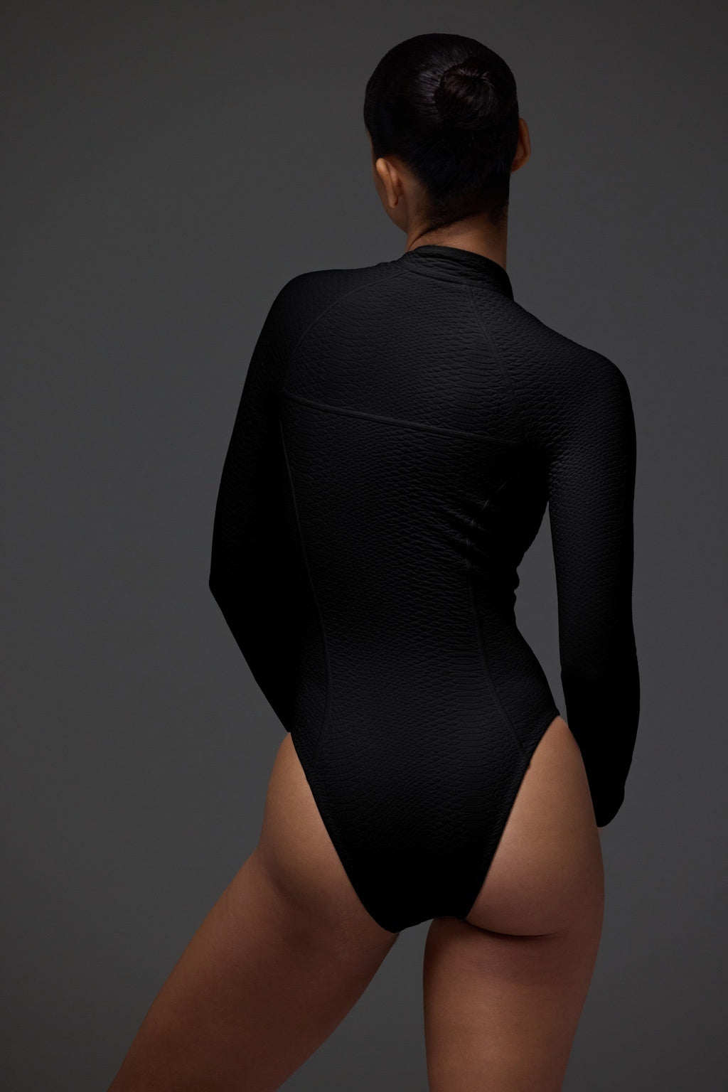 A person with short, dark hair in a bun faces away, wearing the textured black Sophie Surfsuit by Gigi C Bikinis, made from snakeskin embossed fabric, against a dark gray background.