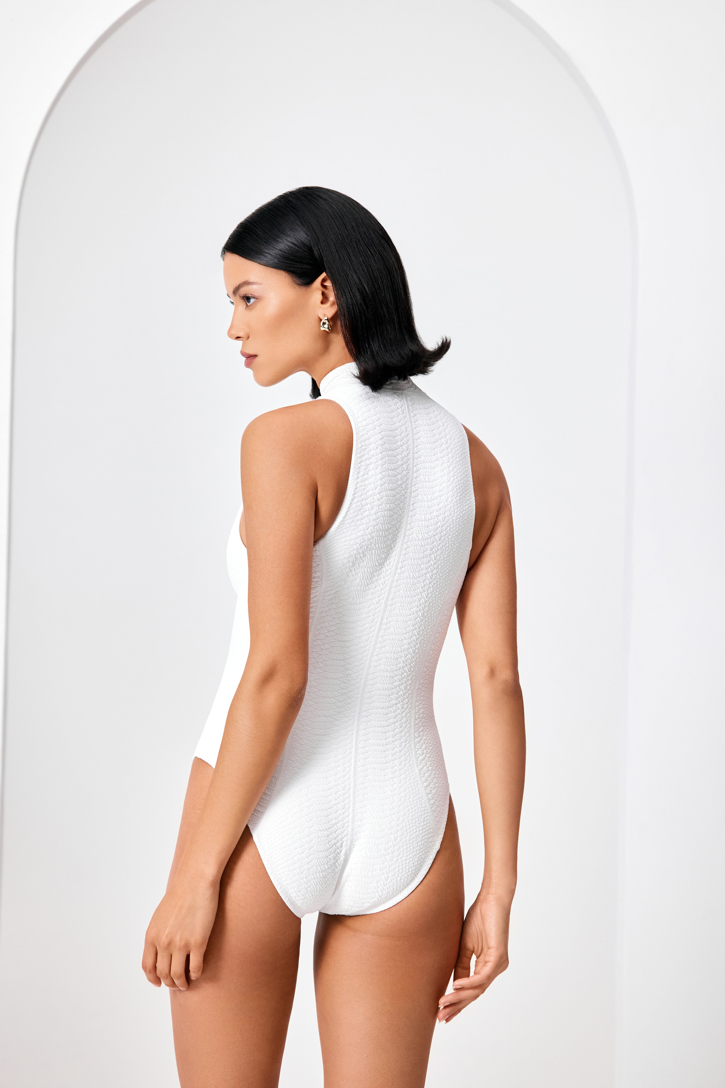 A dark-haired individual is wearing the sleek Roxanne One Piece by Gigi C Bikinis, standing turned away against a minimalist arched white backdrop.