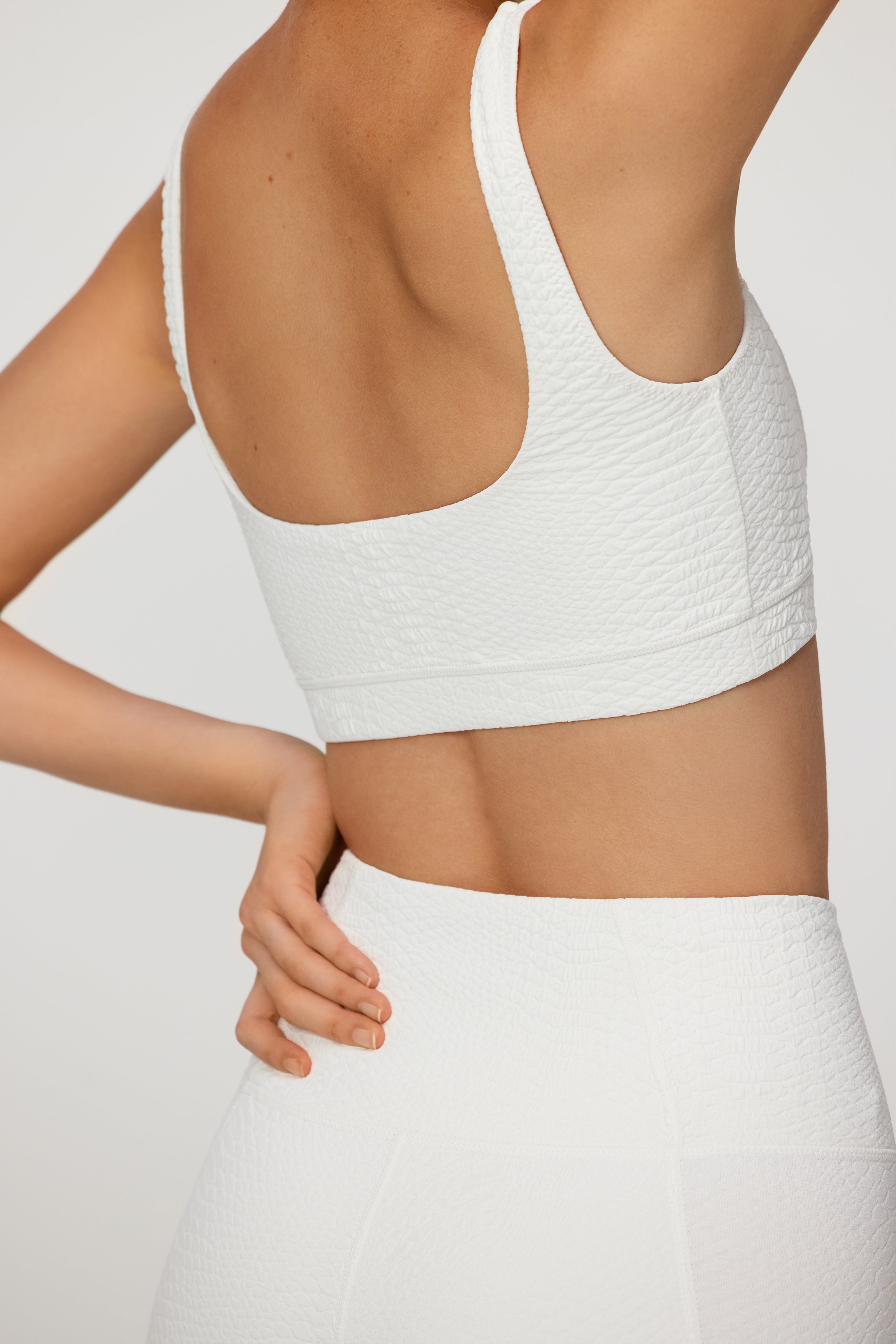 In a minimalist setting, a person elegantly poses with one arm raised and another on their hip, dressed in Gigi C Sports Wilson Bra—a textured white athletic crop top featuring an integrated shelf bra—paired with matching high-waisted leggings.