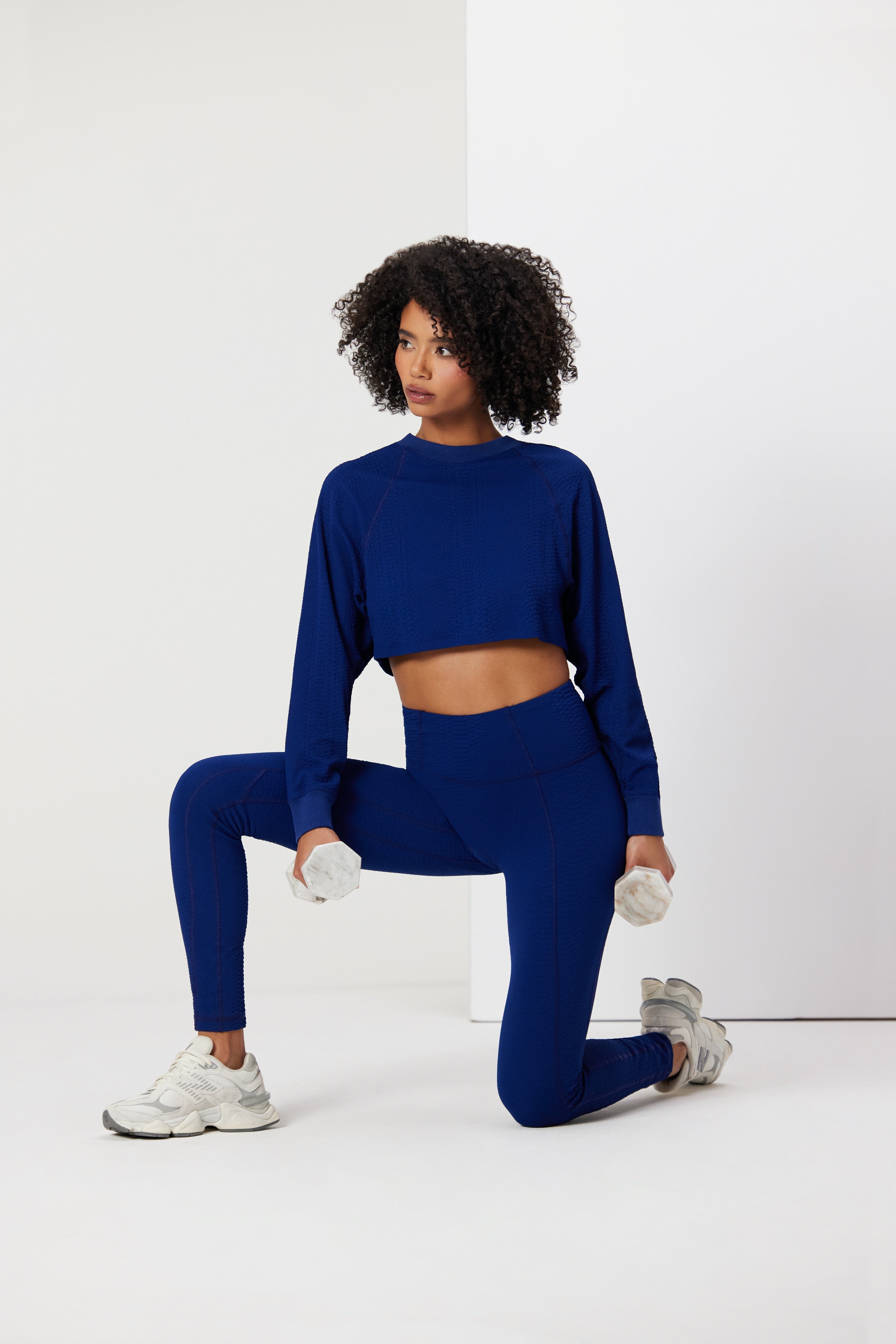 A person in blue workout gear kneels on one knee, holding small weights and wearing a Gigi C Sport Isabella Pullover with white sneakers, posed against a minimalist white background.