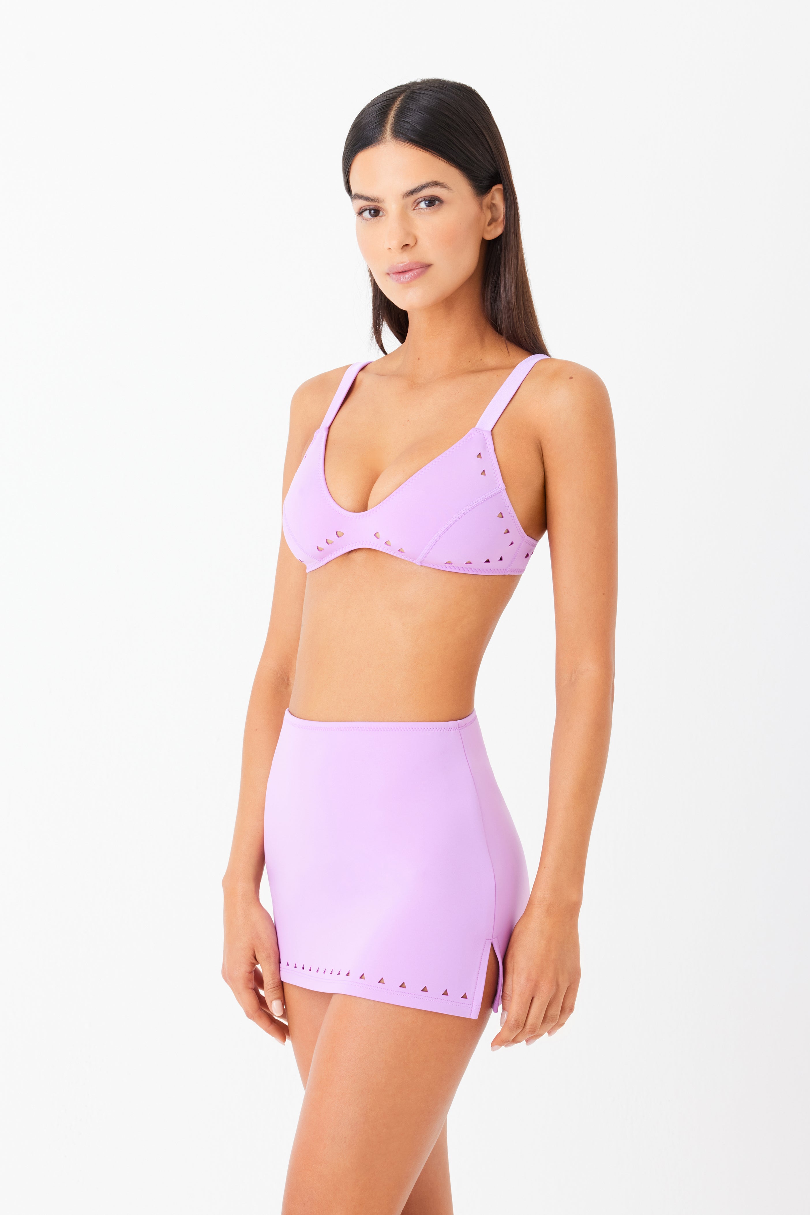 A woman models the Gigi C Bikinis Abby Skirt, a matching lavender mini skirt with intricate laser cutouts, paired with a bikini top. She poses against a white background, gazing at the camera with her arms by her sides and long hair cascading down.