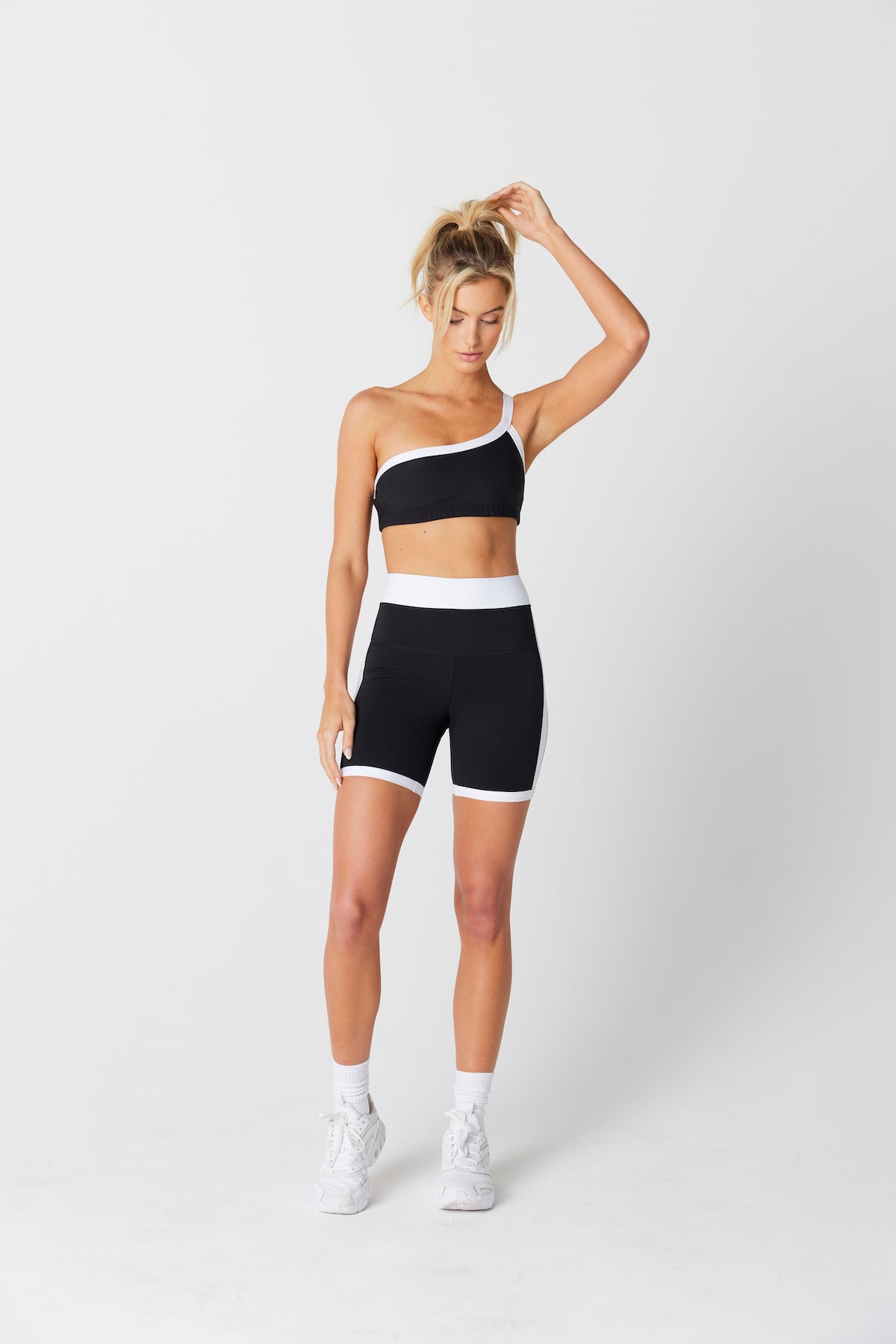 A woman poses against a plain backdrop in the Gigi C Sport Addie Asymmetric Bra, made of breathable knit fabric, paired with matching shorts. Her left arm touches her high ponytail as she pairs the look with white socks and sneakers.