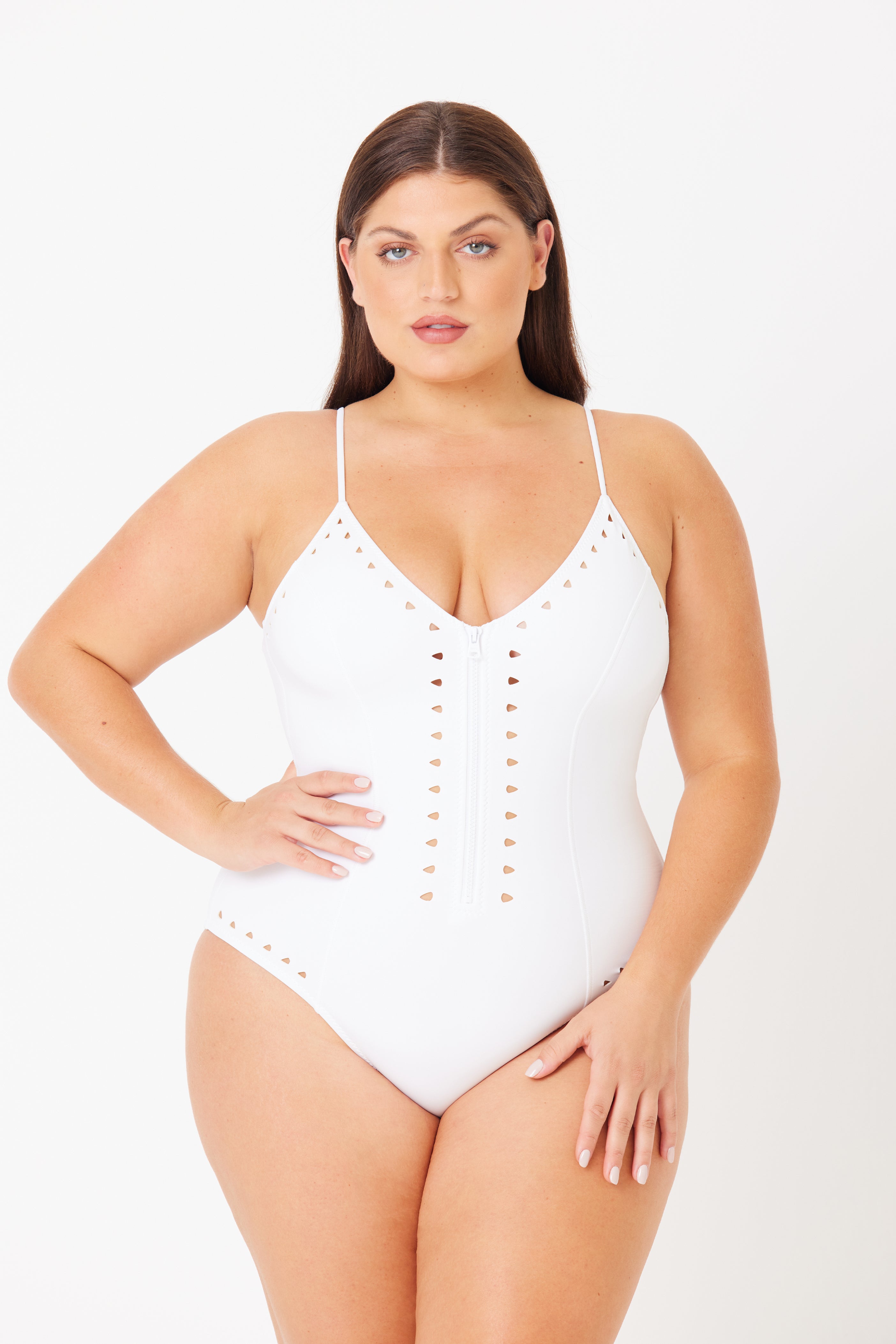 A woman poses confidently in a Gigi C Bikinis Alexandra One Piece Full Coverage swimsuit. This white V-neck features eyelet detailing, a color-match zipper, thin straps, and sleek cutouts, as she stands against a plain white background with one hand on her hip.