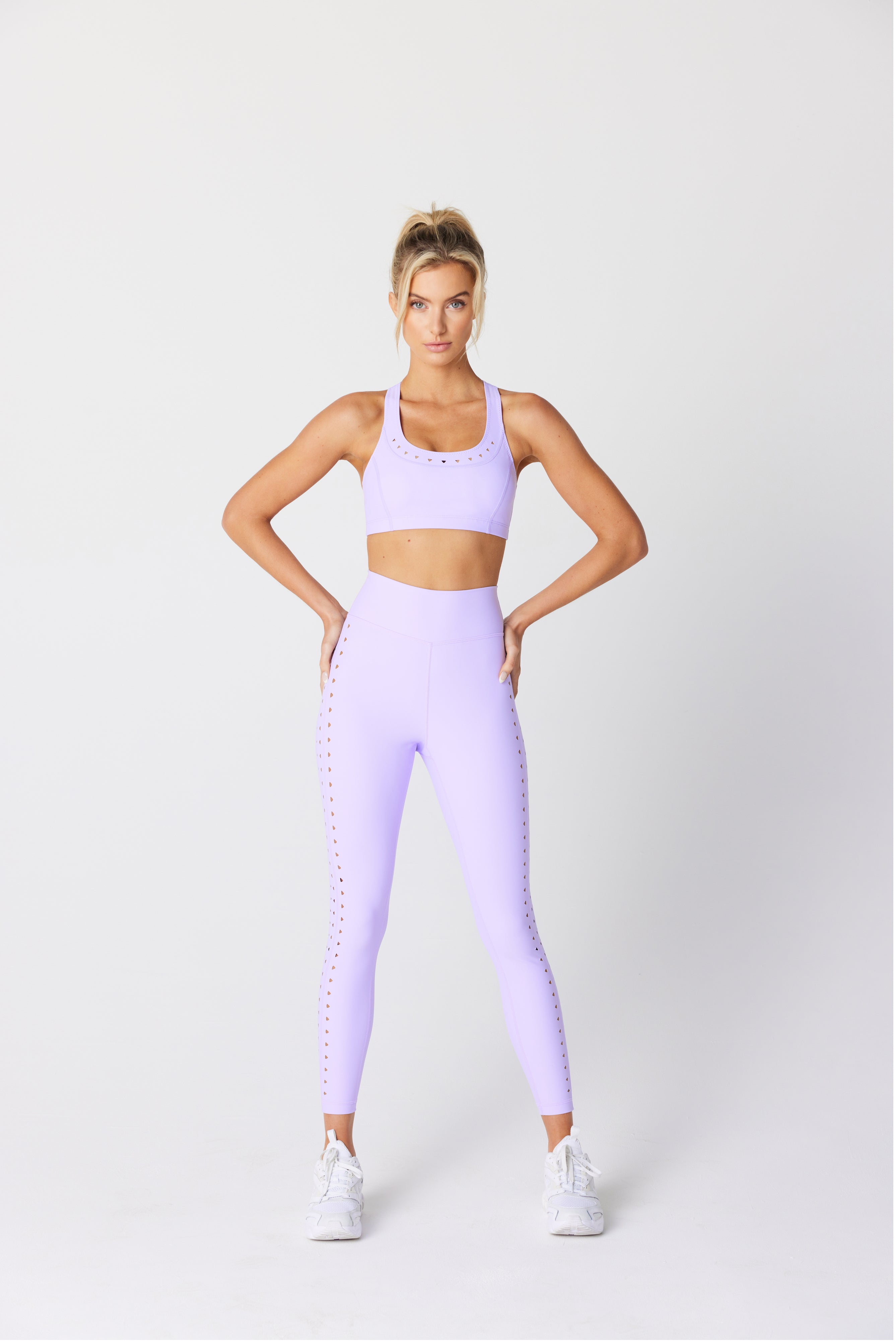 A confident person poses against a white background in Gigi C Sports lavender Annabelle Bra Top and matching recycled bonded jersey leggings, wearing white sneakers with hands on hips and their hair in a high ponytail.