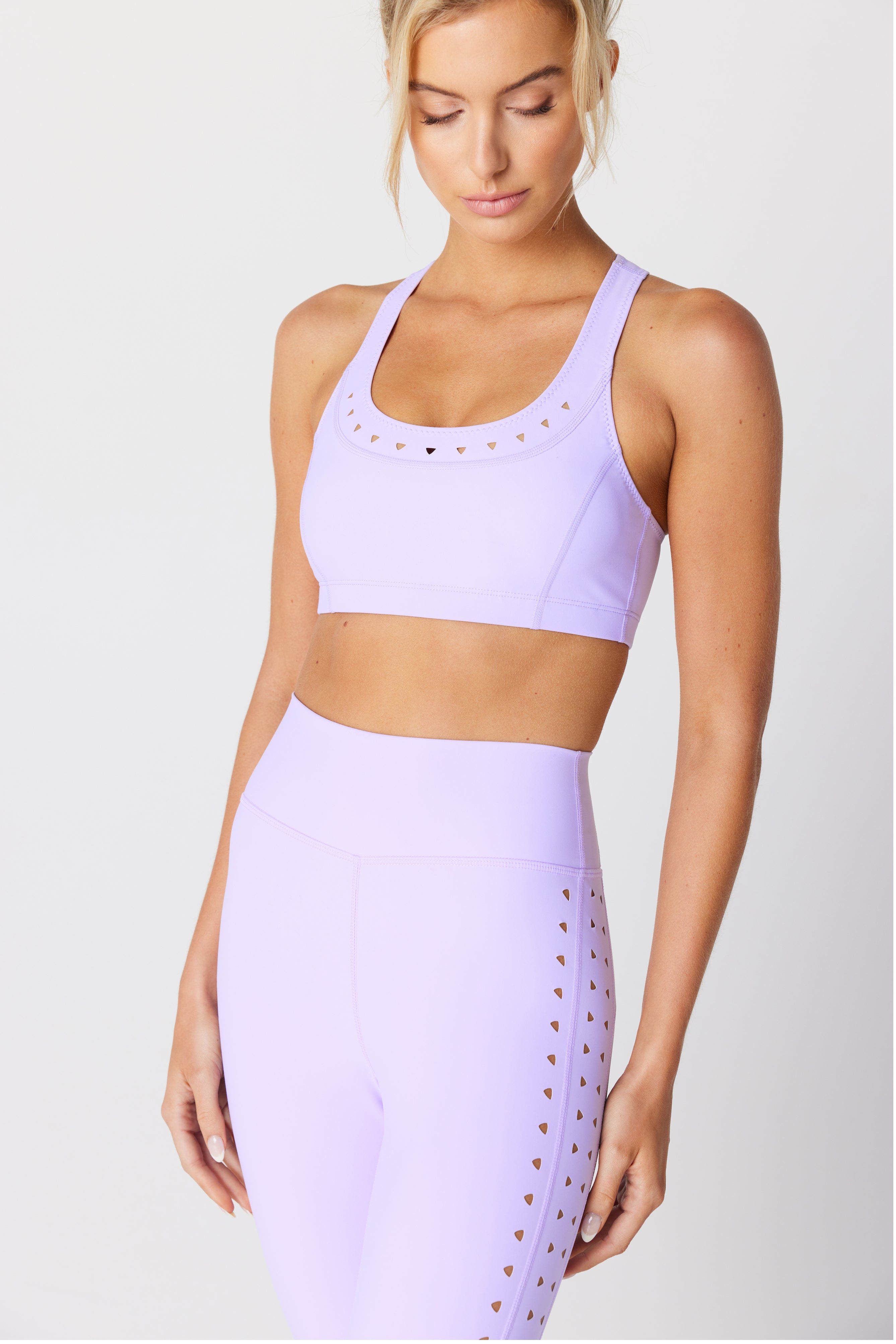 The woman is wearing a Gigi C Sport Annabelle Bra Top in recycled bonded jersey with heart-shaped cutouts, paired with lavender leggings. She stands against a plain background, gazing downward, her hair styled in a loose bun.