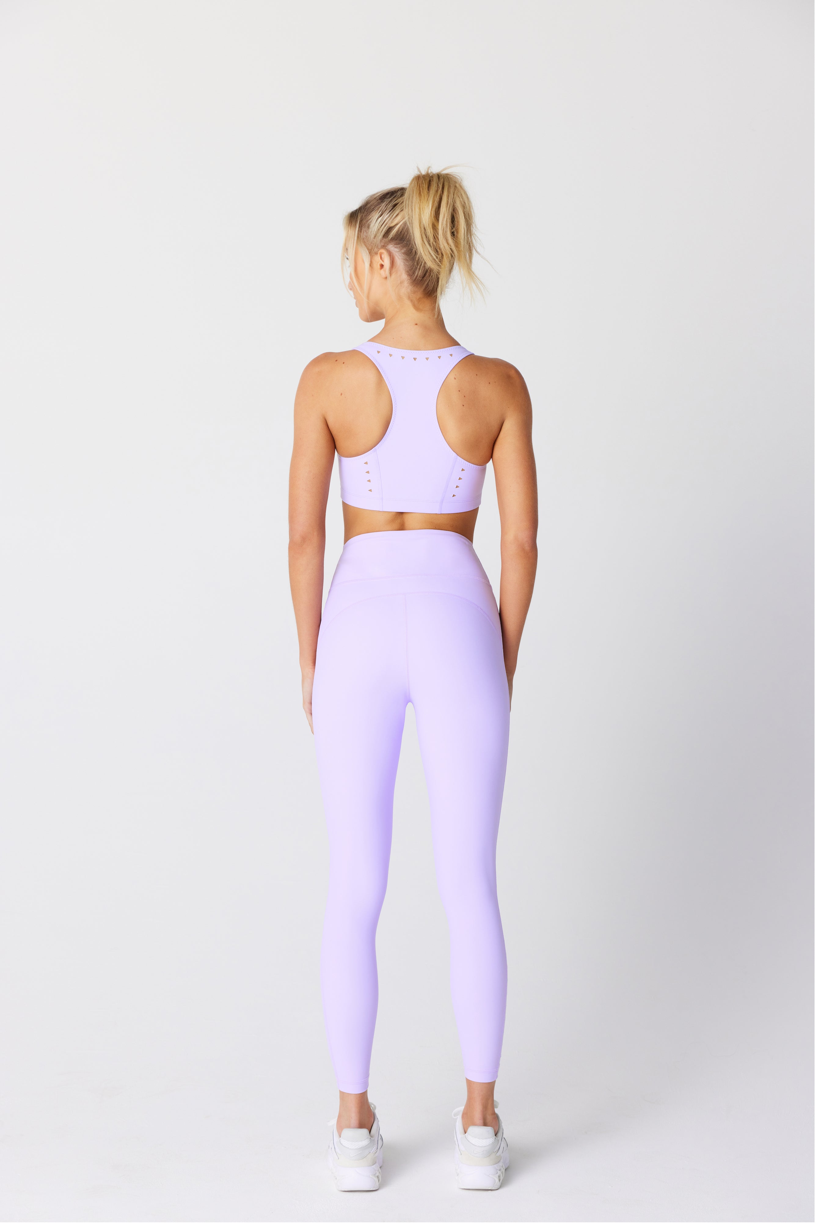 A woman exudes confidence with her back to the camera, wearing a lilac Katrina Legging by Gigi C Sport. Her blonde ponytail accents a high-waisted fit and white sneakers. Made from recycled bonded jersey, her outfit is both stylish and eco-friendly against the plain white backdrop.