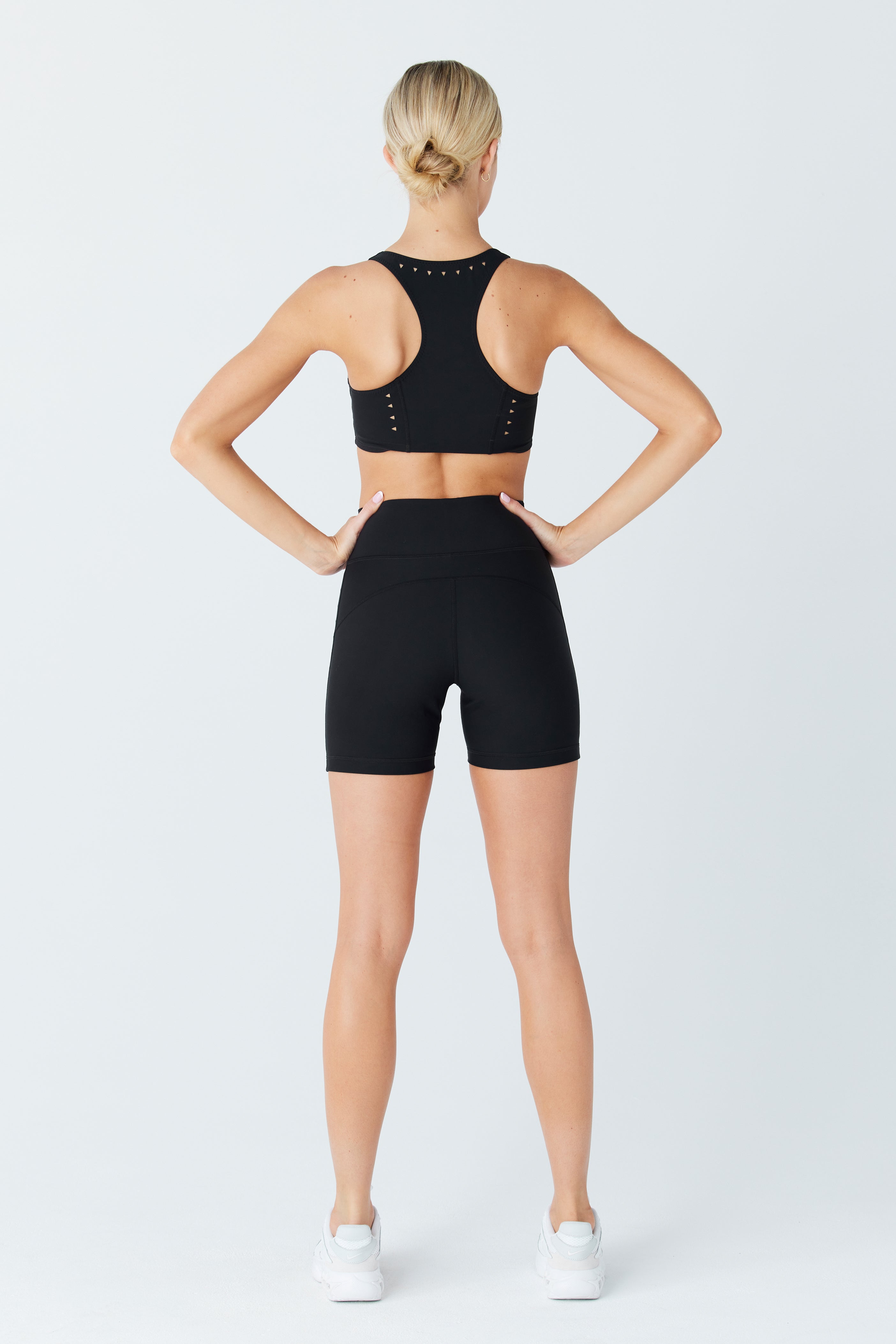 Standing against a light gray background, a person models a black sports bra and Gigi C Sports Hailey Bike Short with laser cutouts, styled with white athletic shoes. Their hair is neatly bunned, emphasizing the ensembles sleek design and compression fabric for an active lifestyle.