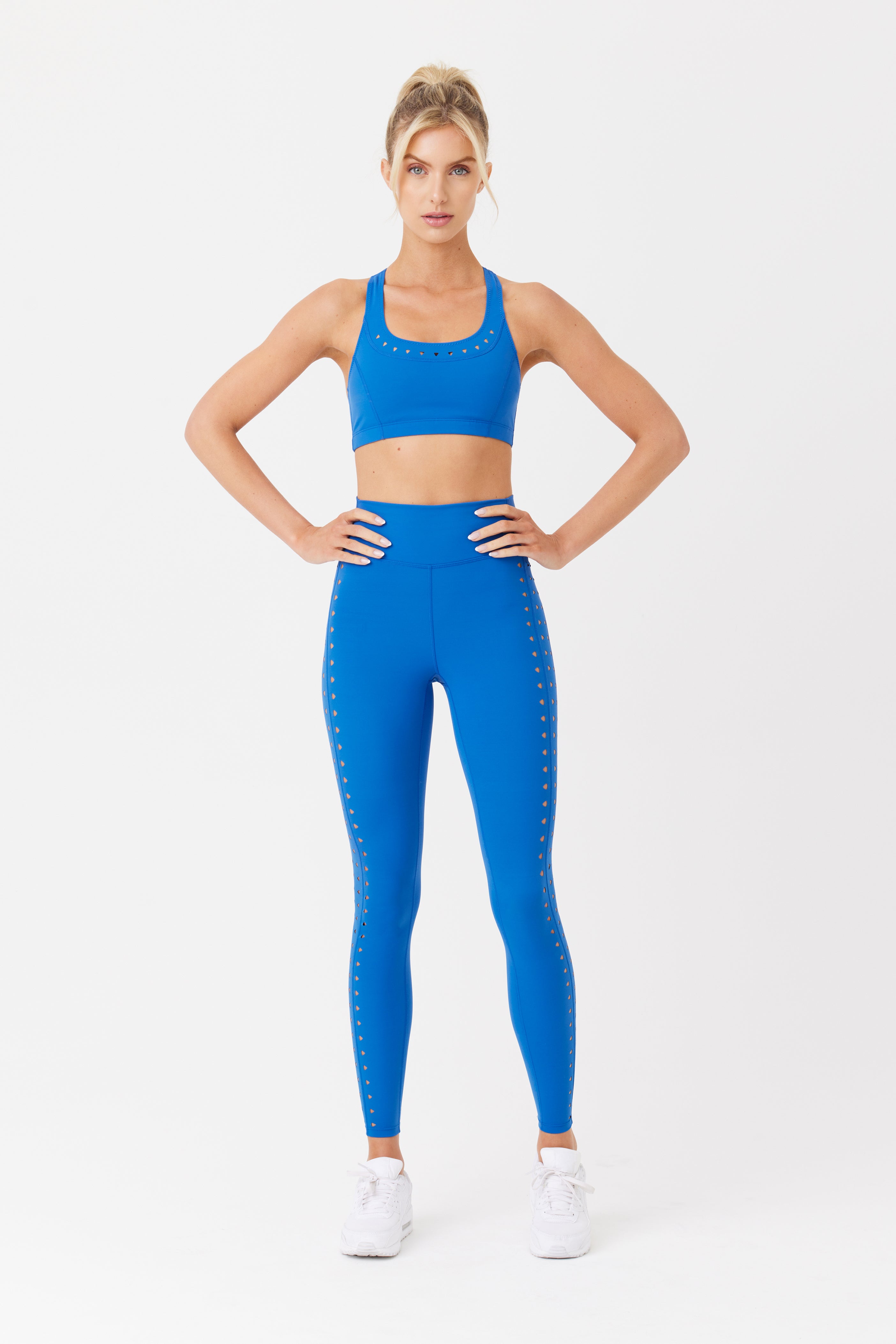 Wearing the Gigi C Sport Annabelle Bra Top and matching leggings, a person stands confidently with hands on hips. The blue outfit features small decorative elements and is made of breathable fabric. They pair it with white sneakers and a ponytail against a white background.