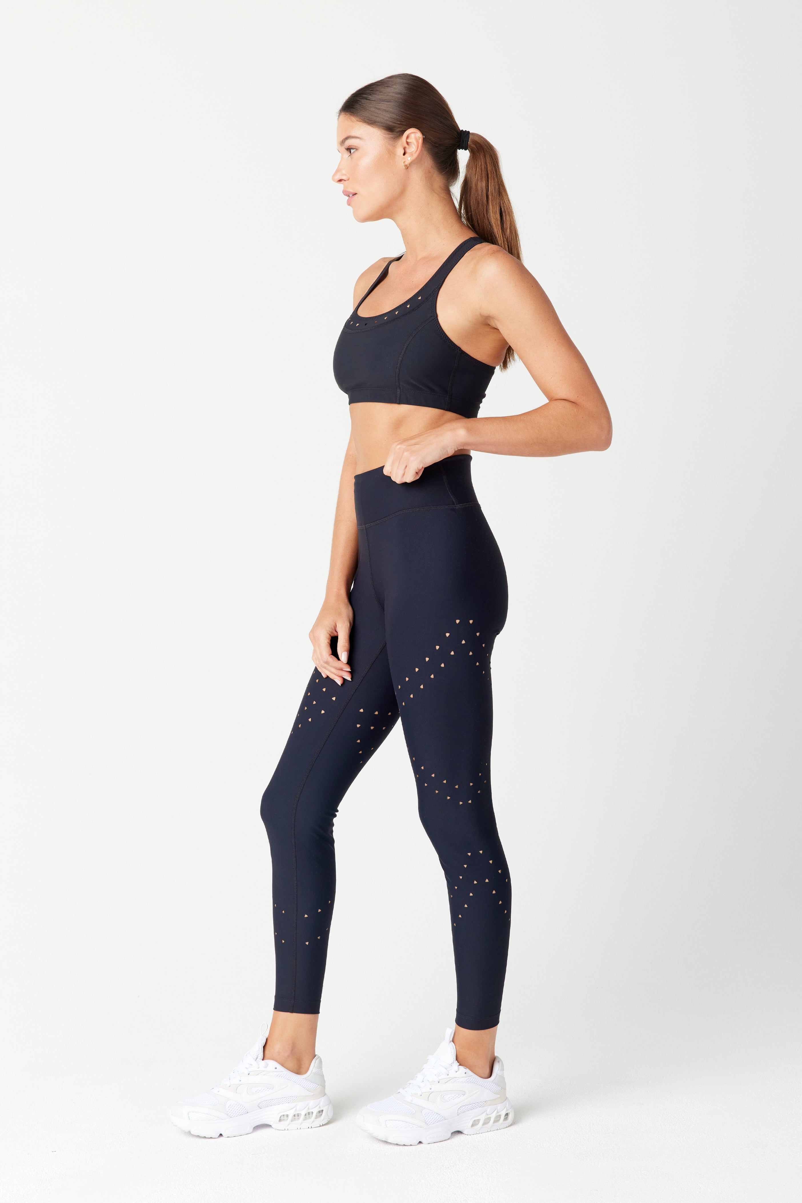 A woman in a black Gigi C Sport Annabelle Bra Top and leggings with small perforations poses in profile against a plain background. The outfit, crafted from recycled fabric, pairs perfectly with white sneakers. Her hair in a ponytail and left hand on her hip completes the sleek look.