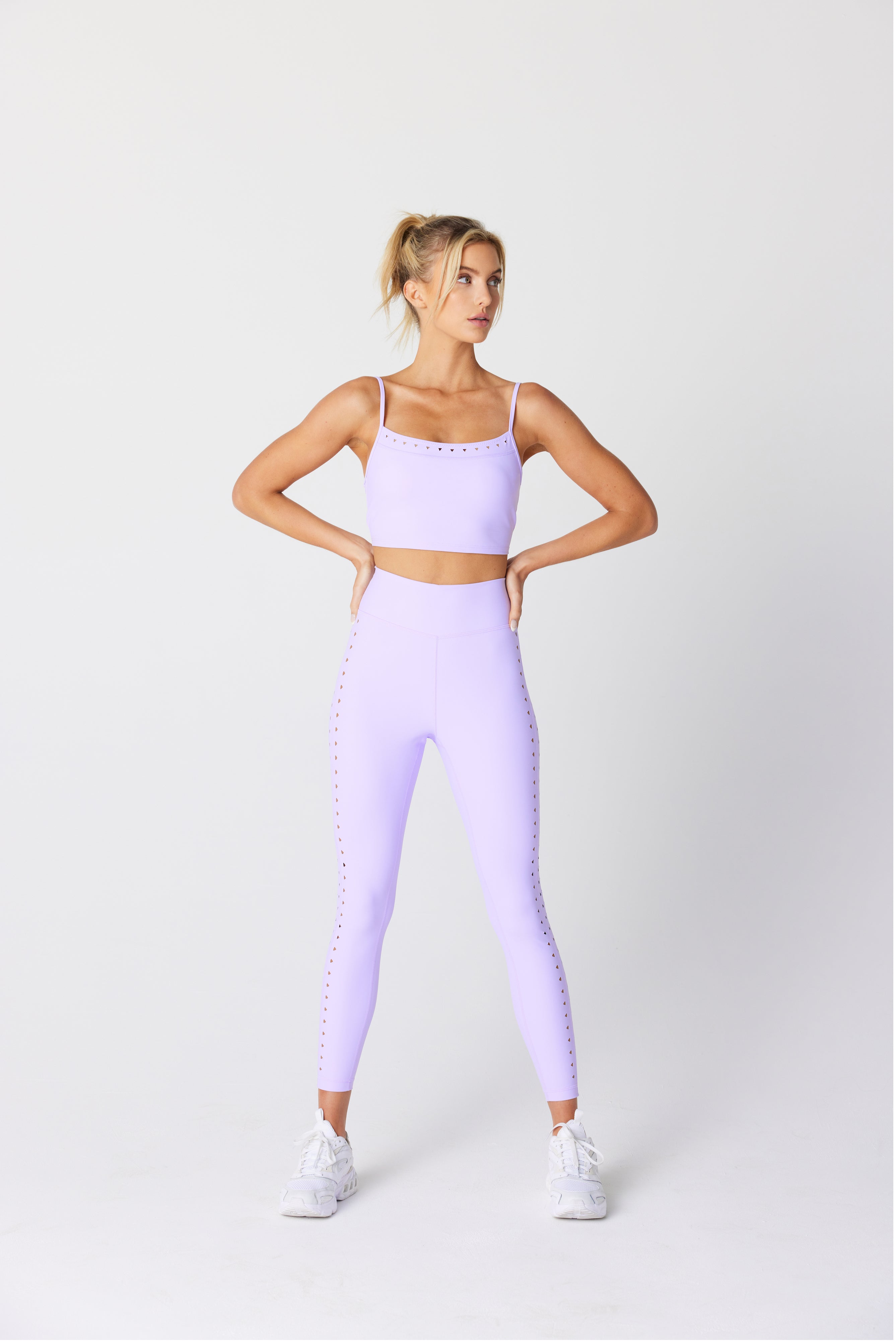 A person in Gigi C Sports Avie Top, a light purple workout outfit with breathable fabric and laser cutouts, stands hands on hips against a white background. Theyre wearing white sneakers and have their hair in a ponytail.