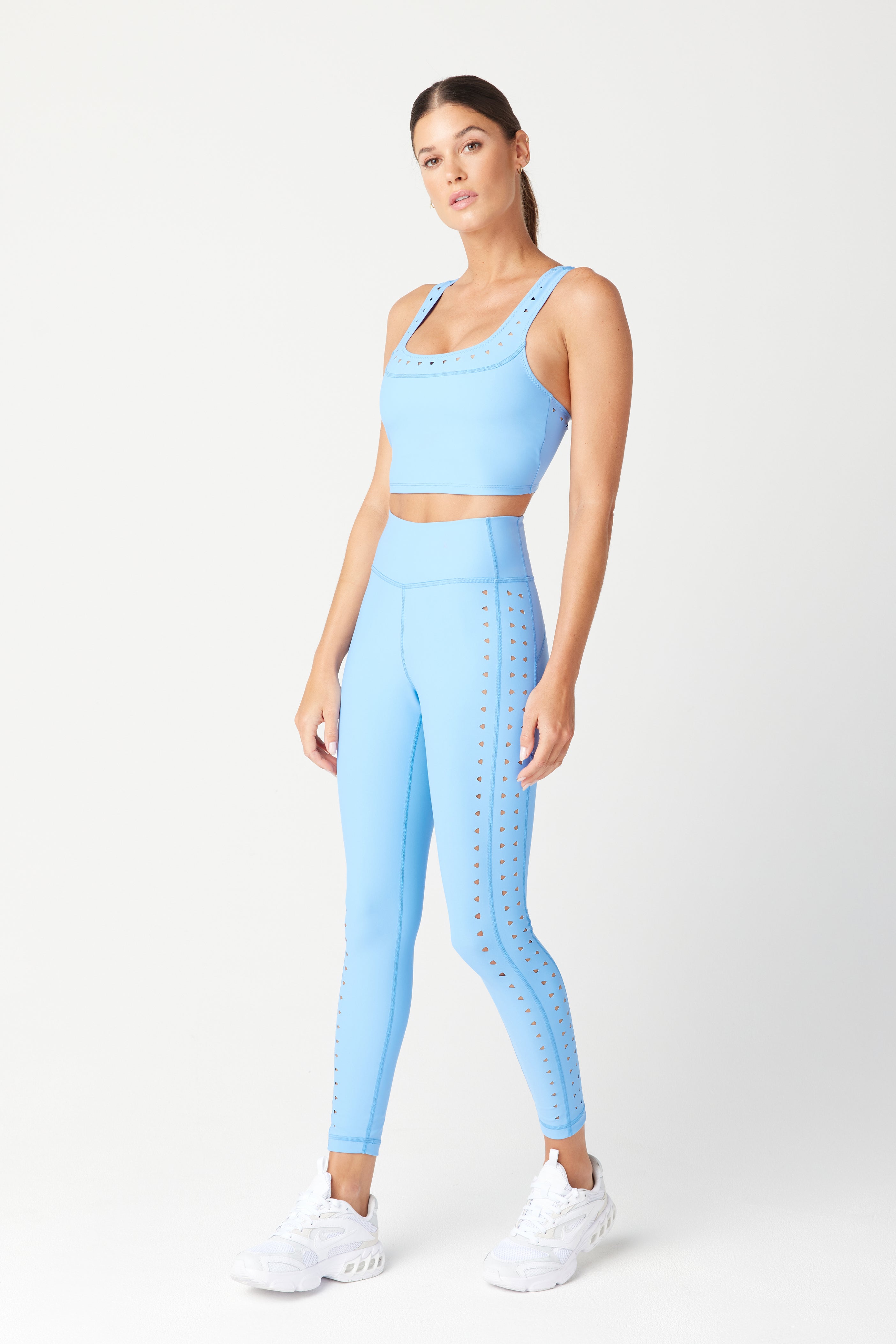 A person stands against a plain backdrop in a light blue Benny Tank by Gigi C Sport and matching high-waisted leggings with triangle cutouts. Made from breathable fabric, they wear white athletic shoes and have their hair tied back, looking slightly to the side.