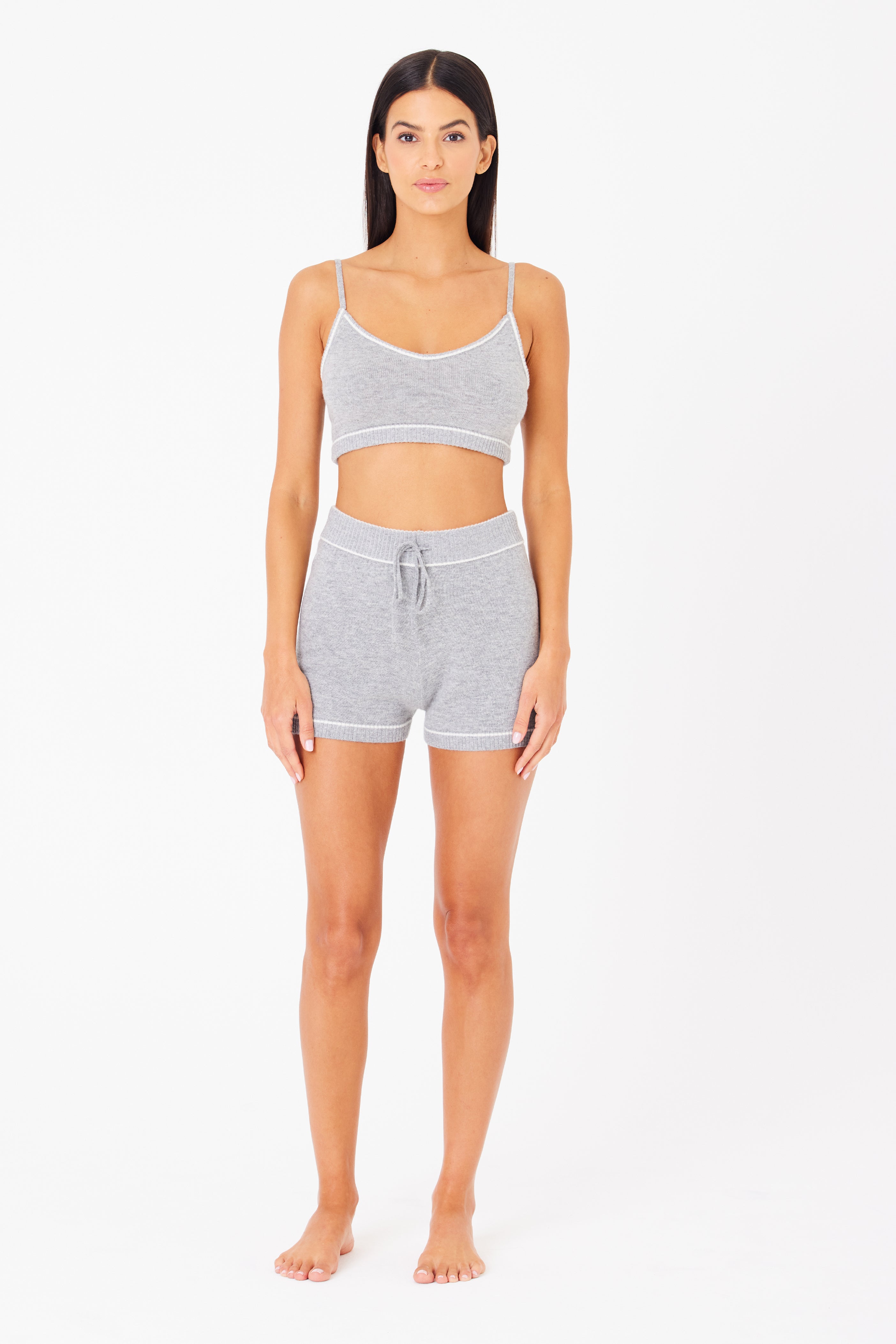 Standing against a white backdrop, they exude casual elegance in a gray Danica Bralette and matching shorts by Gigi C Lounge. With relaxed arms, they embody luxury loungewears epitome—effortless style while barefoot and facing forward.