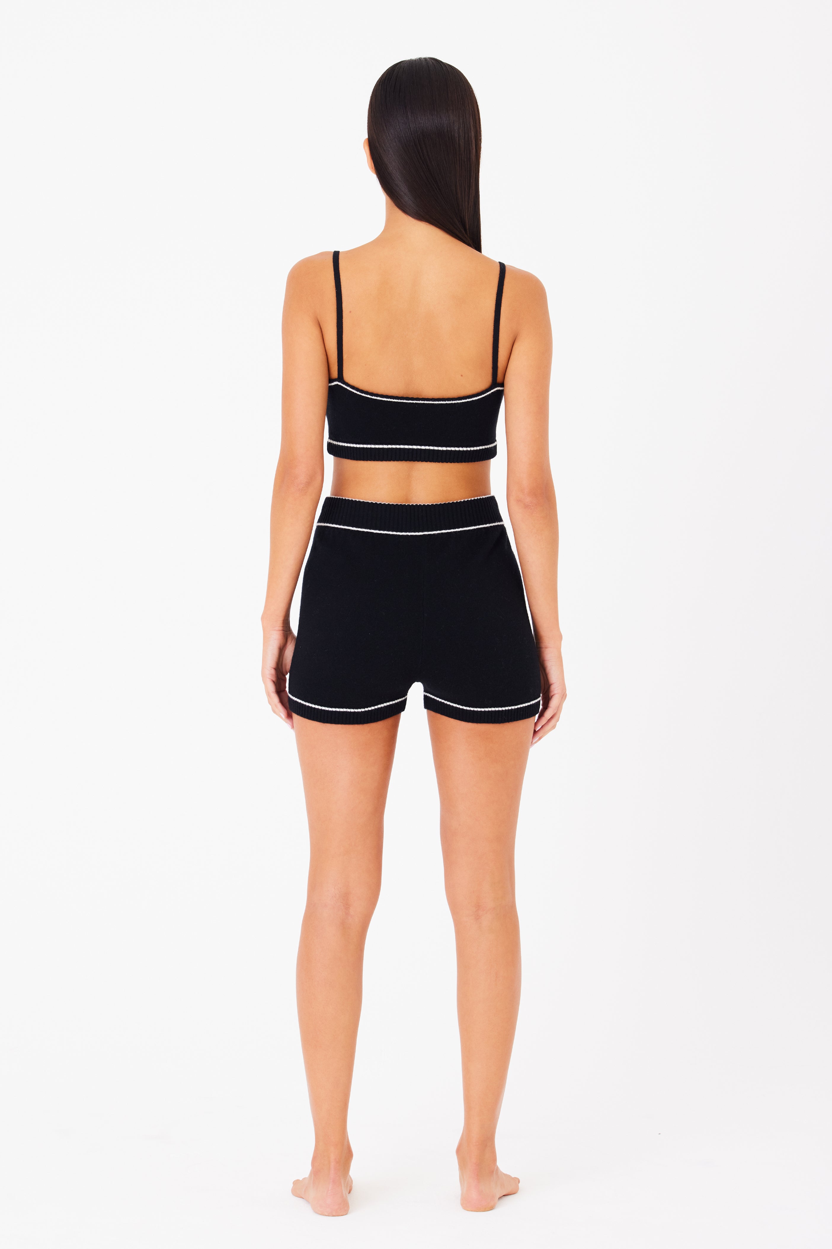 A person with long hair stands barefoot on a white background, facing away from the camera, wearing the Danica Short—a black spaghetti strap crop top and matching black shorts with white trim by Gigi C Lounge, all part of a luxurious cashmere loungewear set.