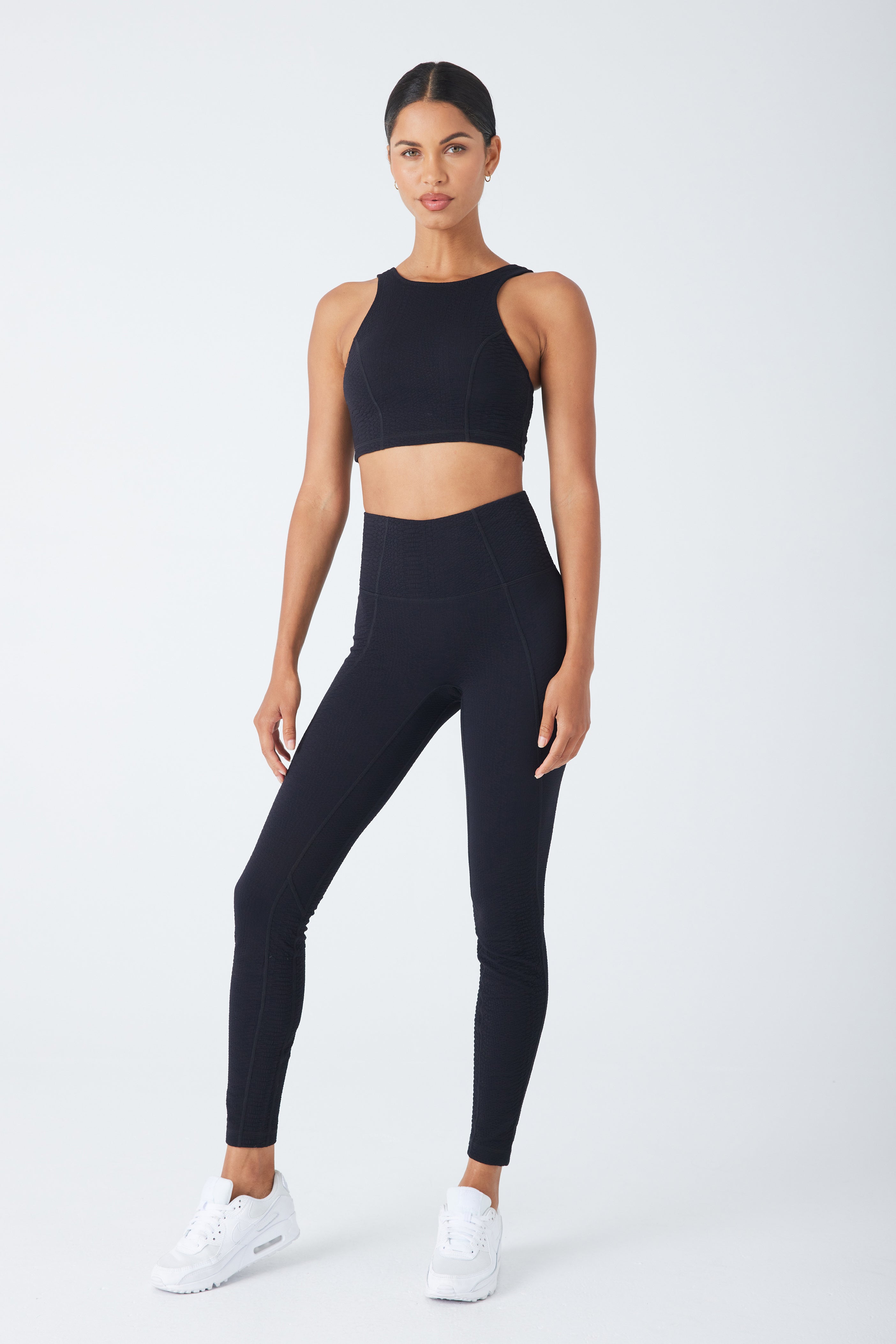 Wearing the Devon Top from Gigi C Sport, a person in a black athletic set, consisting of a medium-support sleeveless crop top and high-waisted bi-stretch compression leggings, pairs the look with white sneakers against a plain white backdrop.