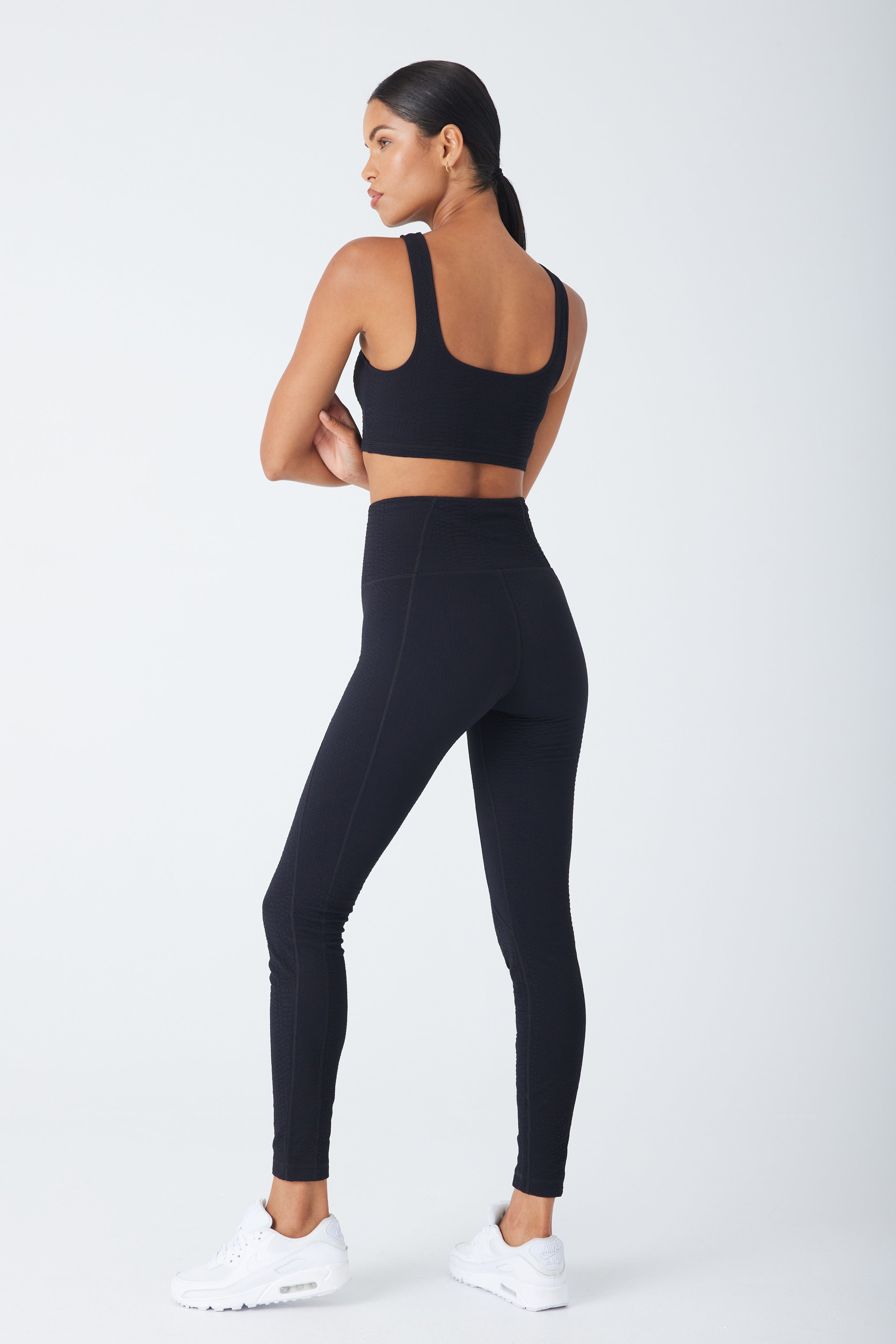 A woman is in a white space wearing the Devon Top by Gigi C Sport, medium support sports bra, leggings, and white athletic shoes. Her neatly tied-back hair and the ensembles sleek bi-stretch compression fit enhance her silhouette as she gazes to the side.