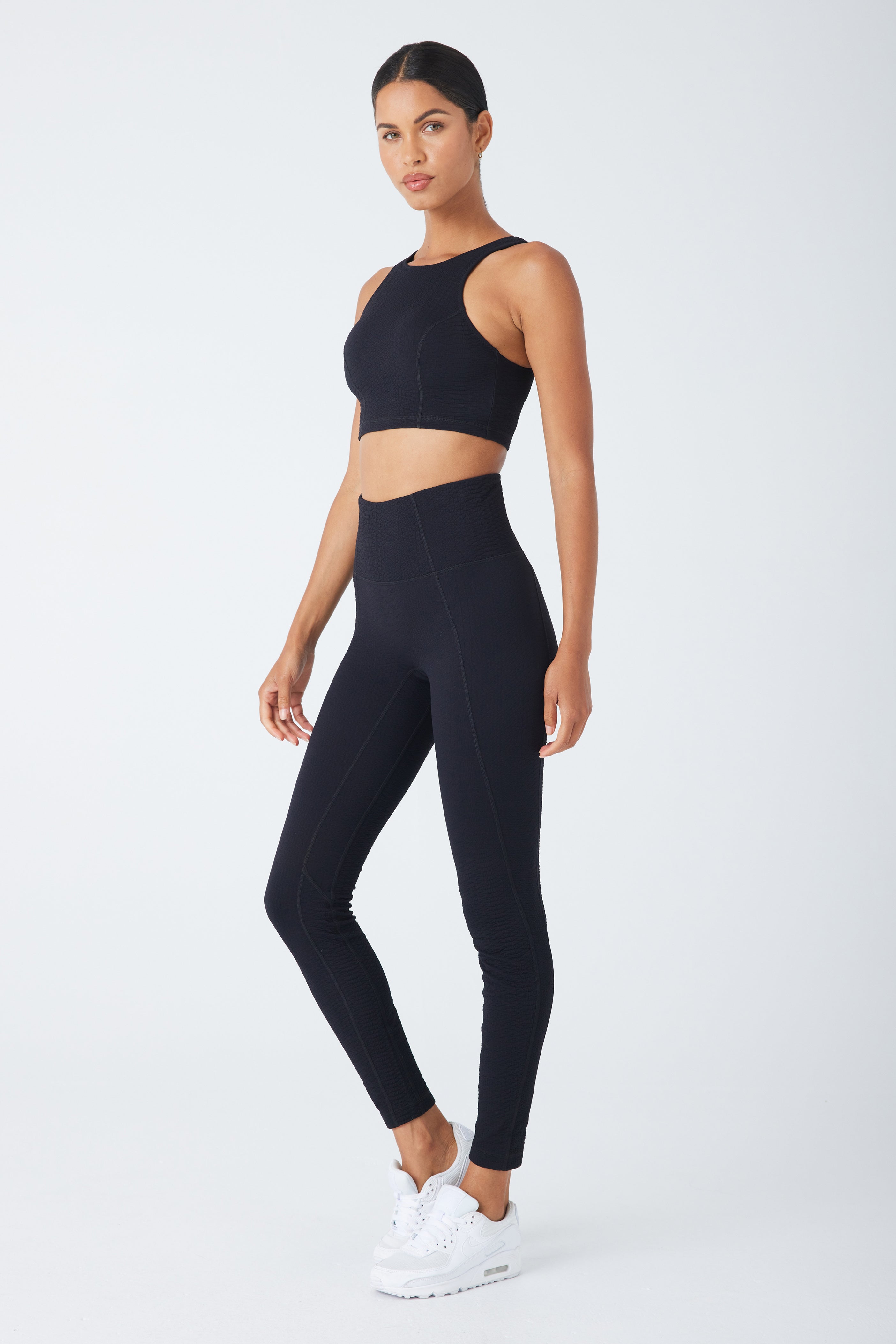 A woman stands against a white backdrop wearing the Devon Top—a black athletic, sleeveless crop from Gigi C Sport—paired with high-waisted bi-stretch compression leggings and sleek white sneakers, confidently gazing at the camera.