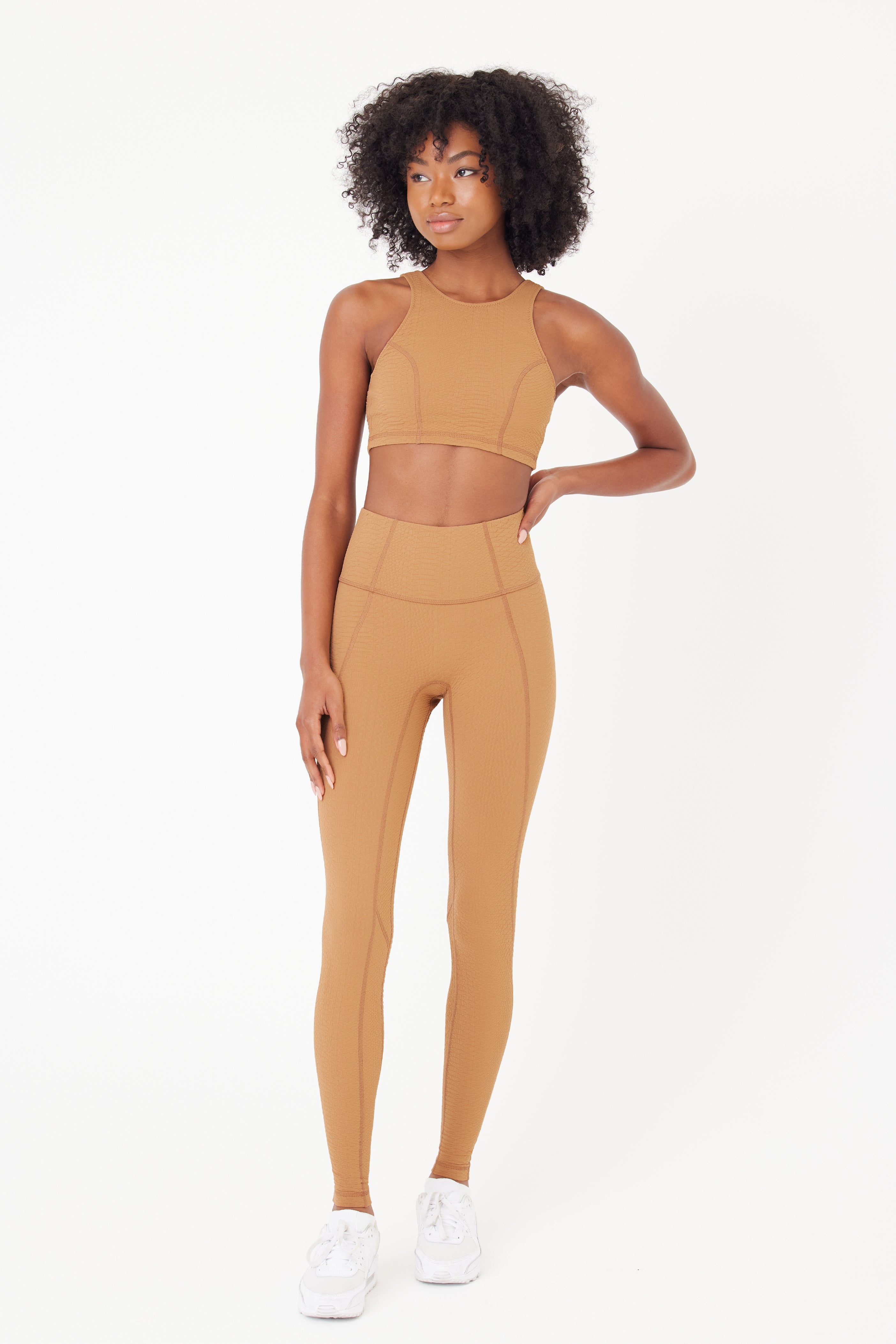 A confident individual stands against a plain white backdrop in a brown Devon Top and matching high-waisted leggings from Gigi C Sport, featuring bi-stretch compression. Curly hair frames their neutral expression as they flaunt white sneakers, embodying both style and function with one hand on their hip.