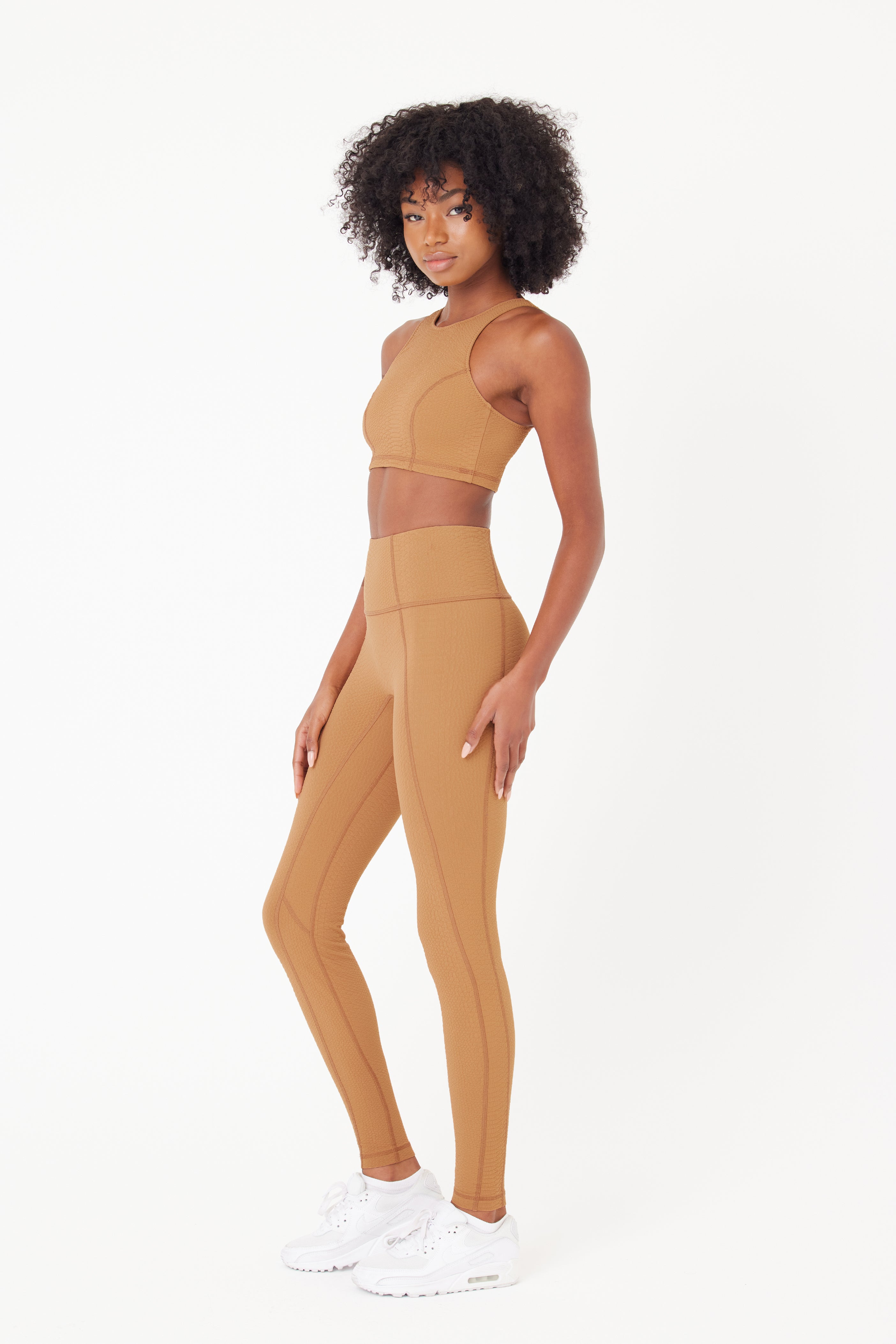 Sporting Gigi C Sports Ava Legging, a curly-haired individual dons a chic brown athletic ensemble of compression waffle jacquard fabric, with a crop top and high-waist leggings. Paired with white sneakers, they stand confidently against a plain white backdrop.