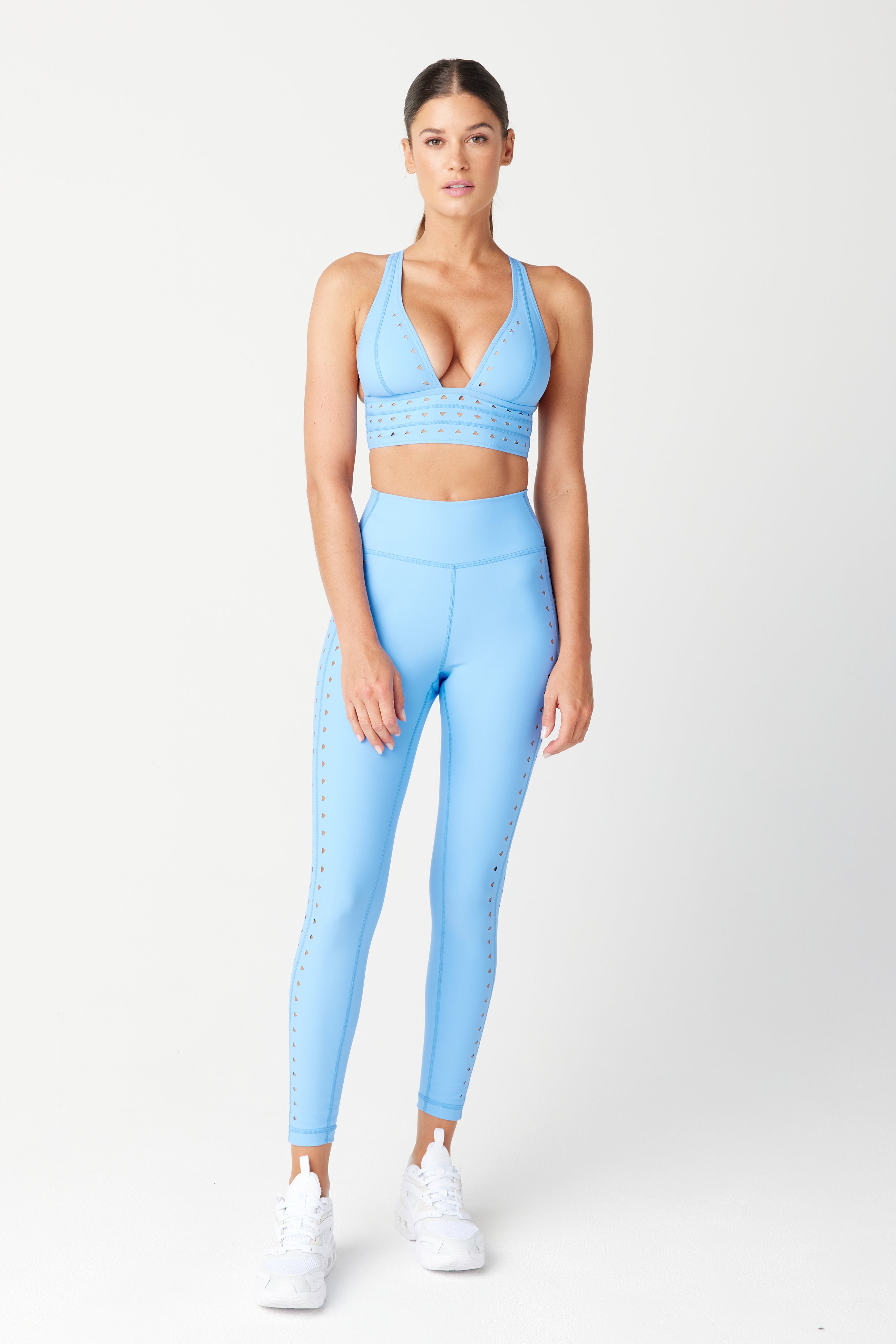 A person stands against a plain background wearing a matching light blue athletic set from Gigi C Sport. The outfit features an Eden Bra with a deep neckline and high-waisted leggings with laser cutouts, all made from recycled bonded jersey, completed with white sneakers.