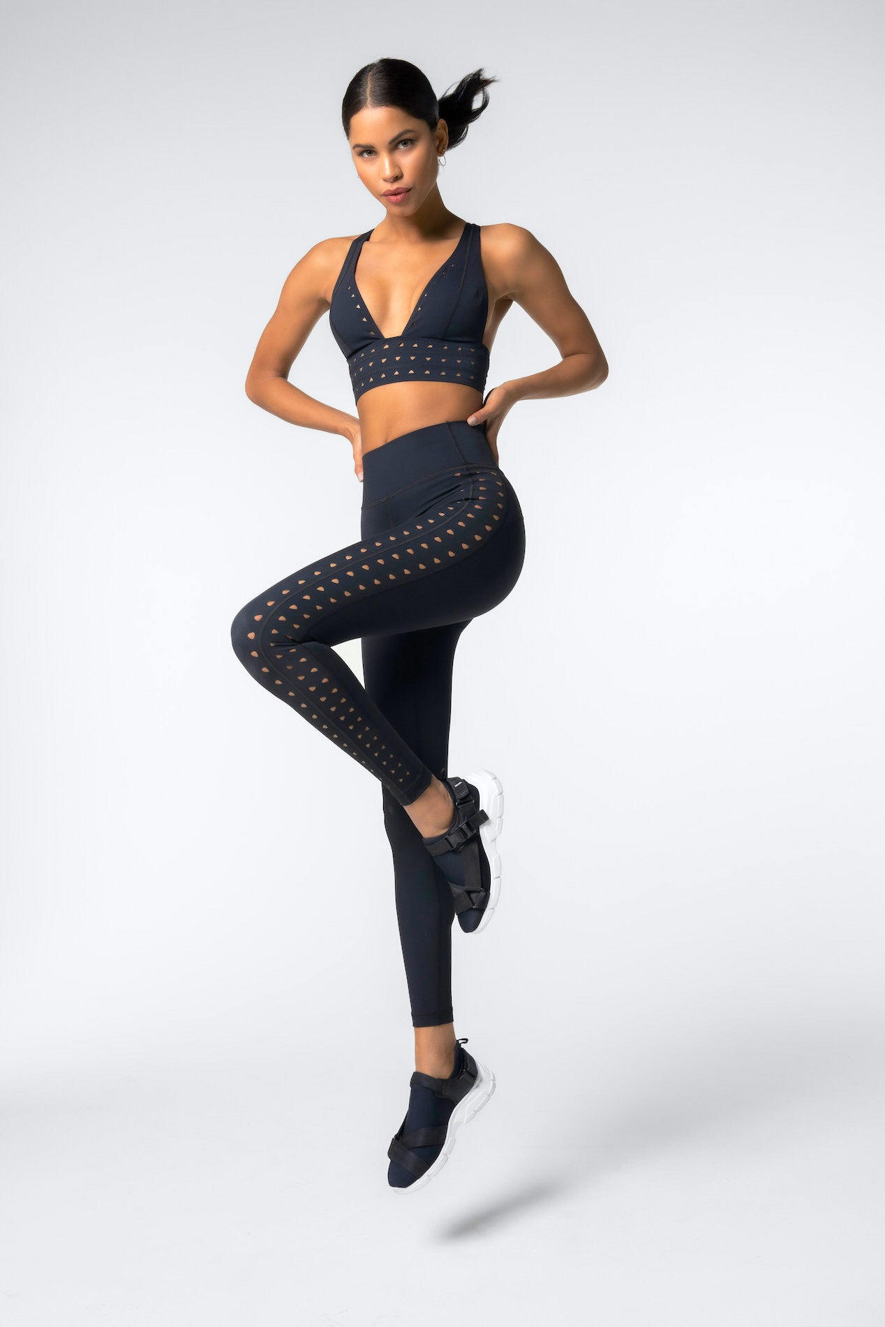 A person wearing a sleek black athletic outfit with laser cutouts, complete with sneakers and Gigi C Sports Eden Bra made from Recycled Bonded Jersey, poses dynamically against a plain backdrop, balancing on one leg with arms akimbo.