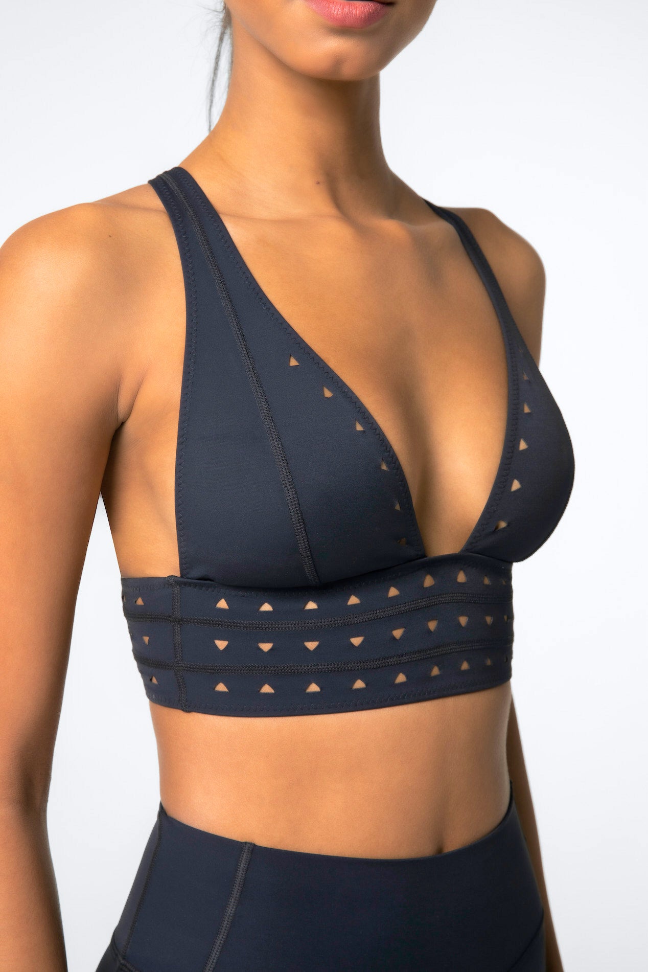 A model in a Gigi C Sport Eden Bra and matching leggings made from Recycled Bonded Jersey, featuring a sleek black design with a deep V-neck and chic laser cutouts, stands against a simple light backdrop.