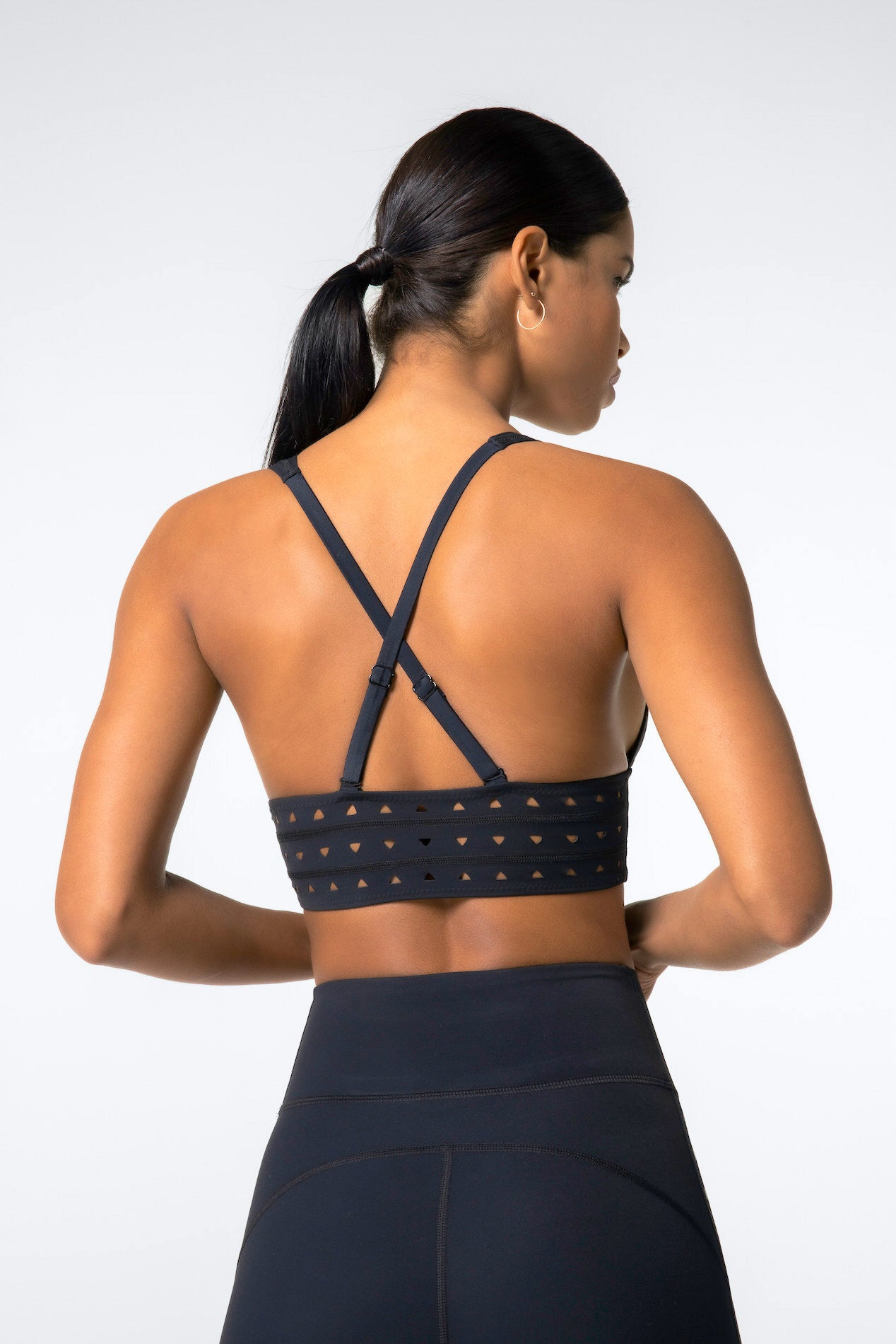 A woman with a ponytail faces away, showcasing Gigi C Sports Eden Bra—black with crisscross straps and laser cutouts—paired with high-waisted black leggings against a plain light gray backdrop.