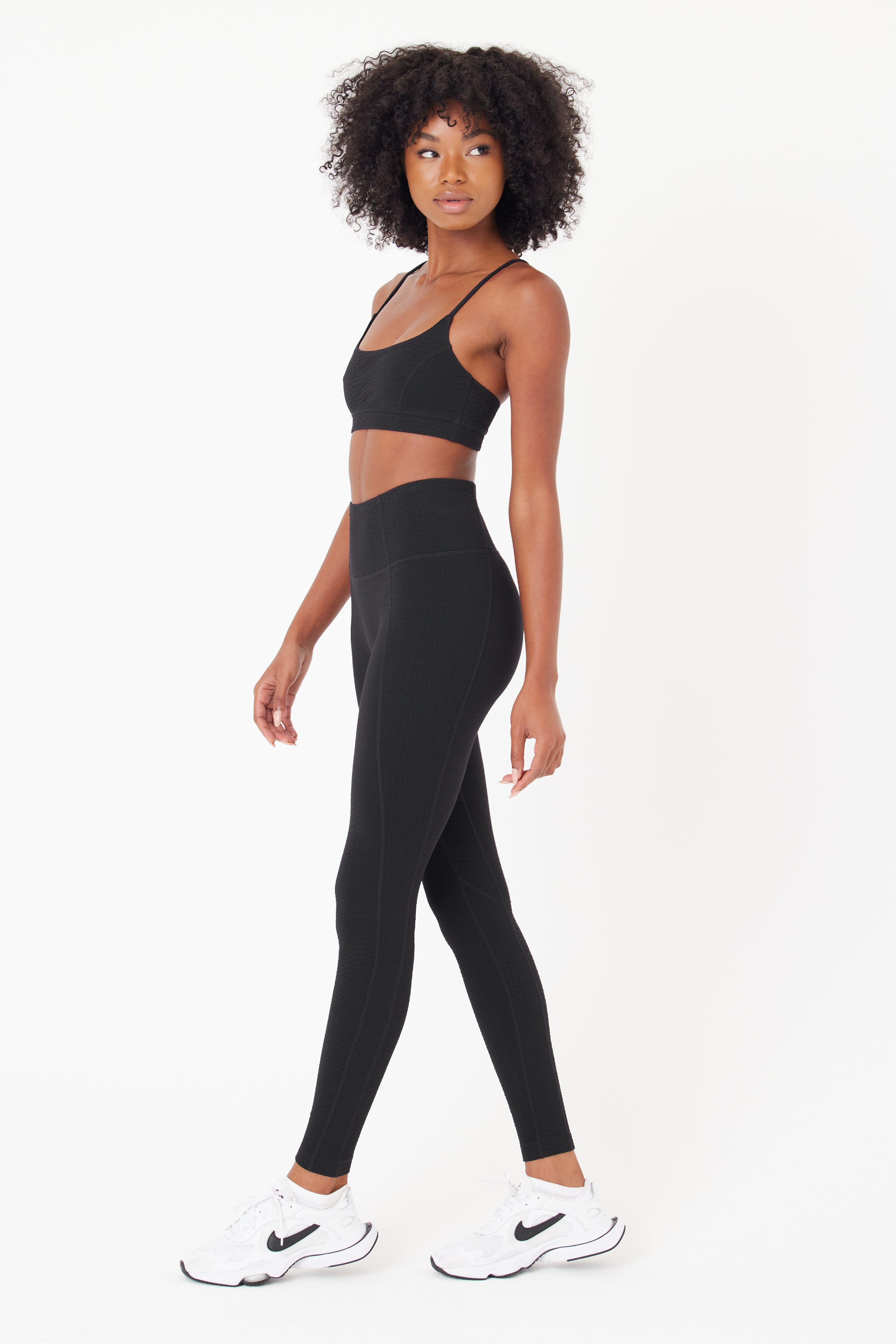 A curly-haired person wears a black sports bra and Gigi C Sports Ava Legging, crafted from bi-stretch compression waffle jacquard fabric, with white sneakers. They stand against a plain white background, facing the camera neutrally.