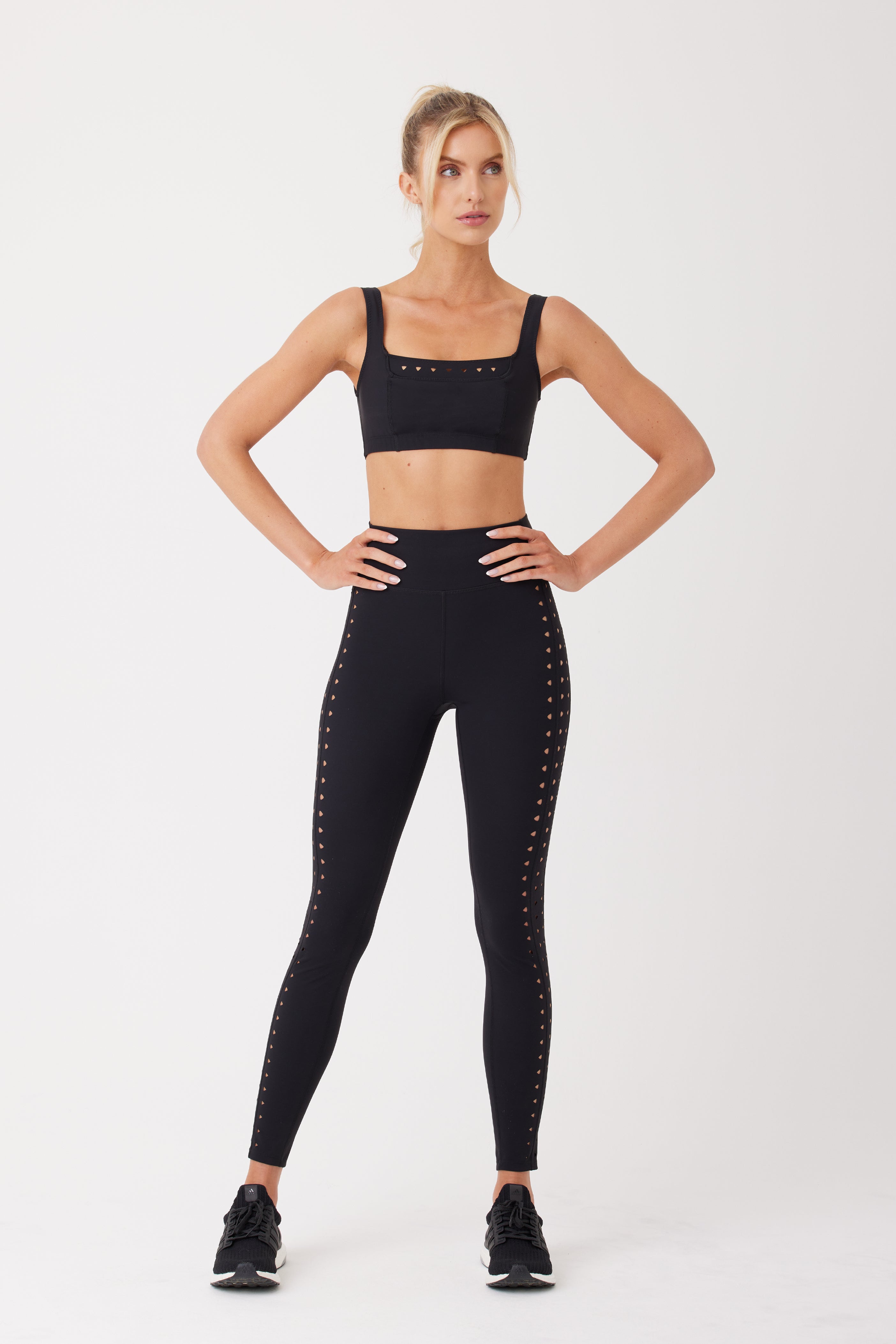 A person poses confidently in a Gigi C Sport Katrina Legging with studded detailing, crafted from compression fabric with laser cutouts, and a black sports bra. Black athletic shoes complete the look against a plain white backdrop.