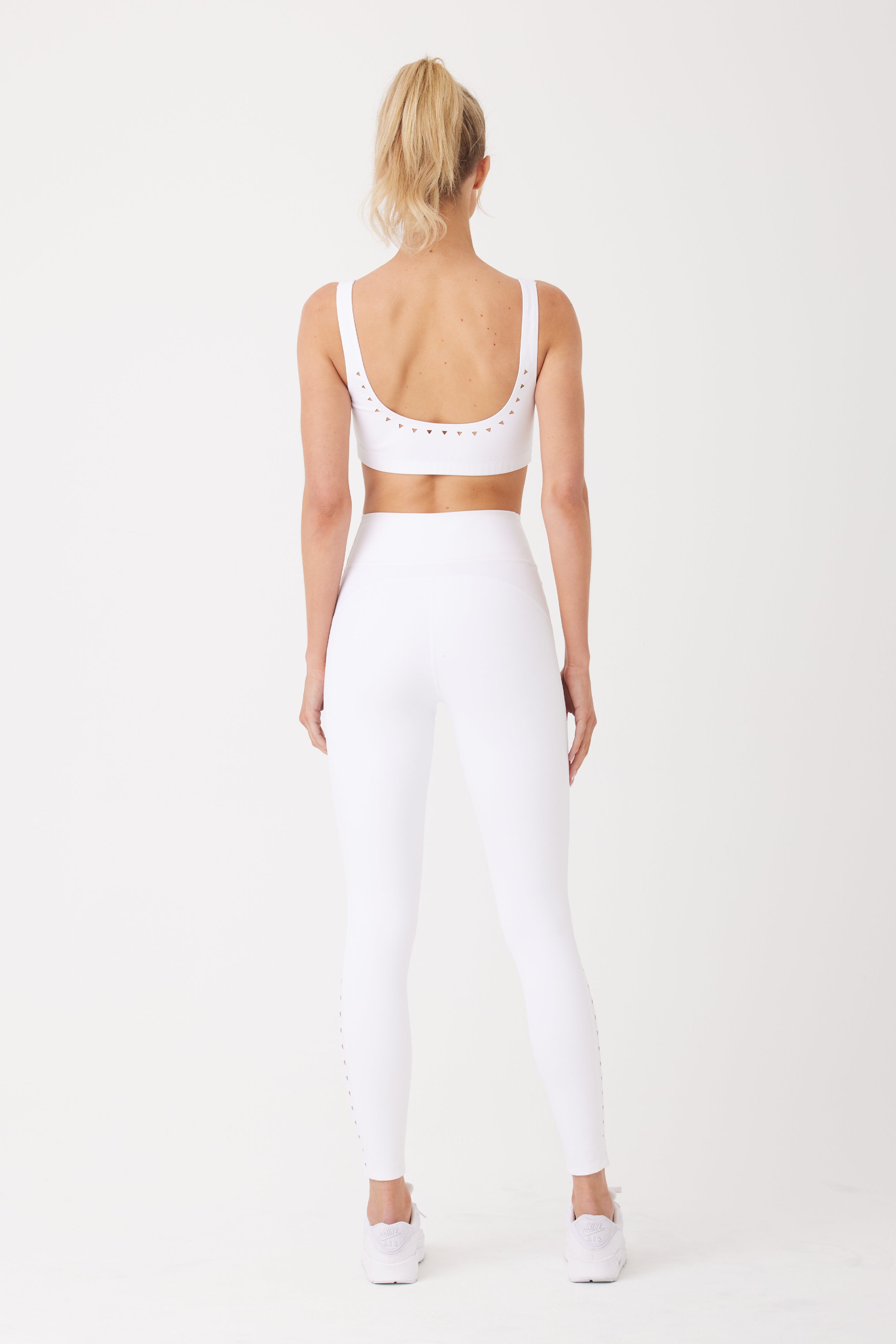 A person with a ponytail faces away, modeling Gigi C Sports Katrina Legging paired with a white sports bra and laser-cut leggings. This sleek ensemble is completed with white sneakers against a plain white background, highlighting sophisticated, modern athletic style.