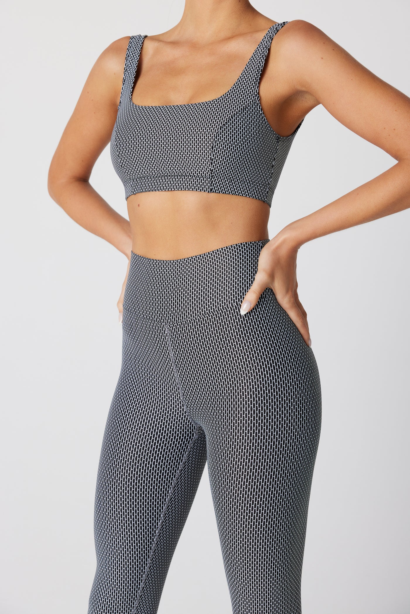 A person wears the Maisie Bra and matching leggings in a chic black and white pattern, crafted from luxurious Italian fabric by Gigi C Sport. Against a pristine white backdrop, they pose with hands on hips to emphasize the sleek design while keeping their face hidden to spotlight the outfit.