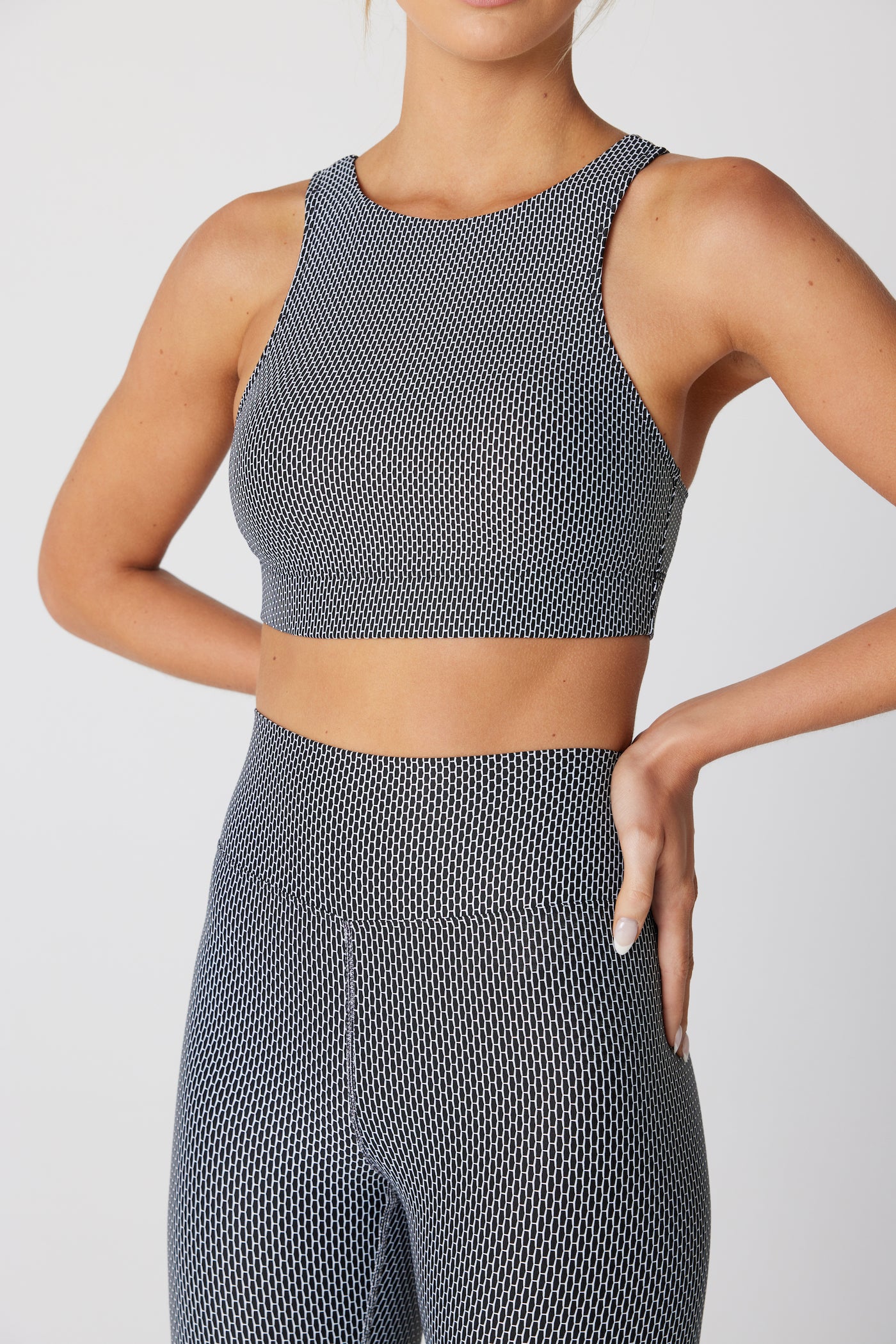 A woman flaunts the Gigi C Sports Nicki Top, a gray and white honeycomb jacquard crop top made of exquisite Italian fabric, paired with matching leggings, as she stands confidently, one hand on her hip against a light background.