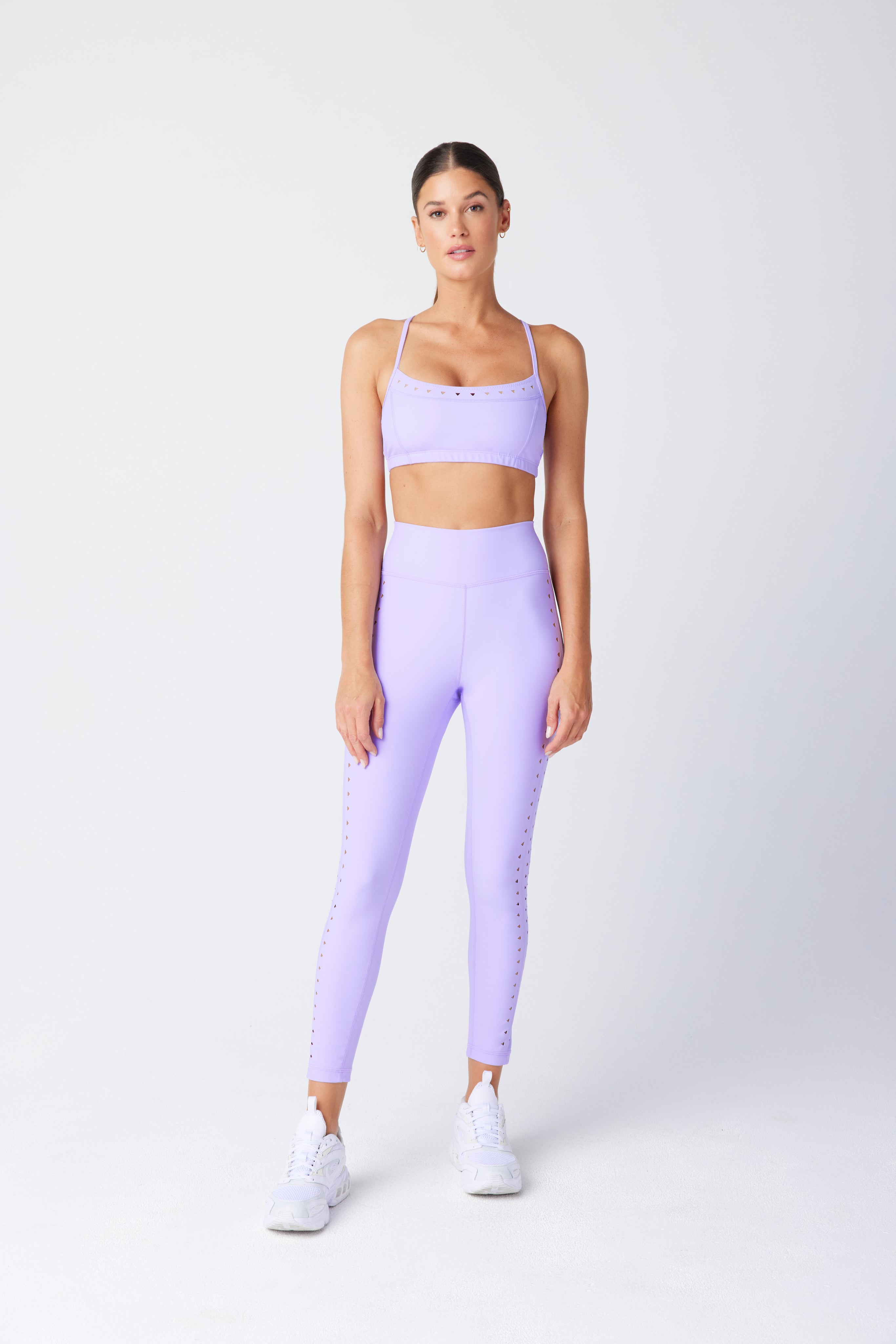 Dressed in a matching lavender workout outfit, featuring the Giselle Bra by Gigi C Sport with adjustable straps and recycled fabric high-waisted leggings, complemented by white sneakers, they stand against a plain light-colored background.