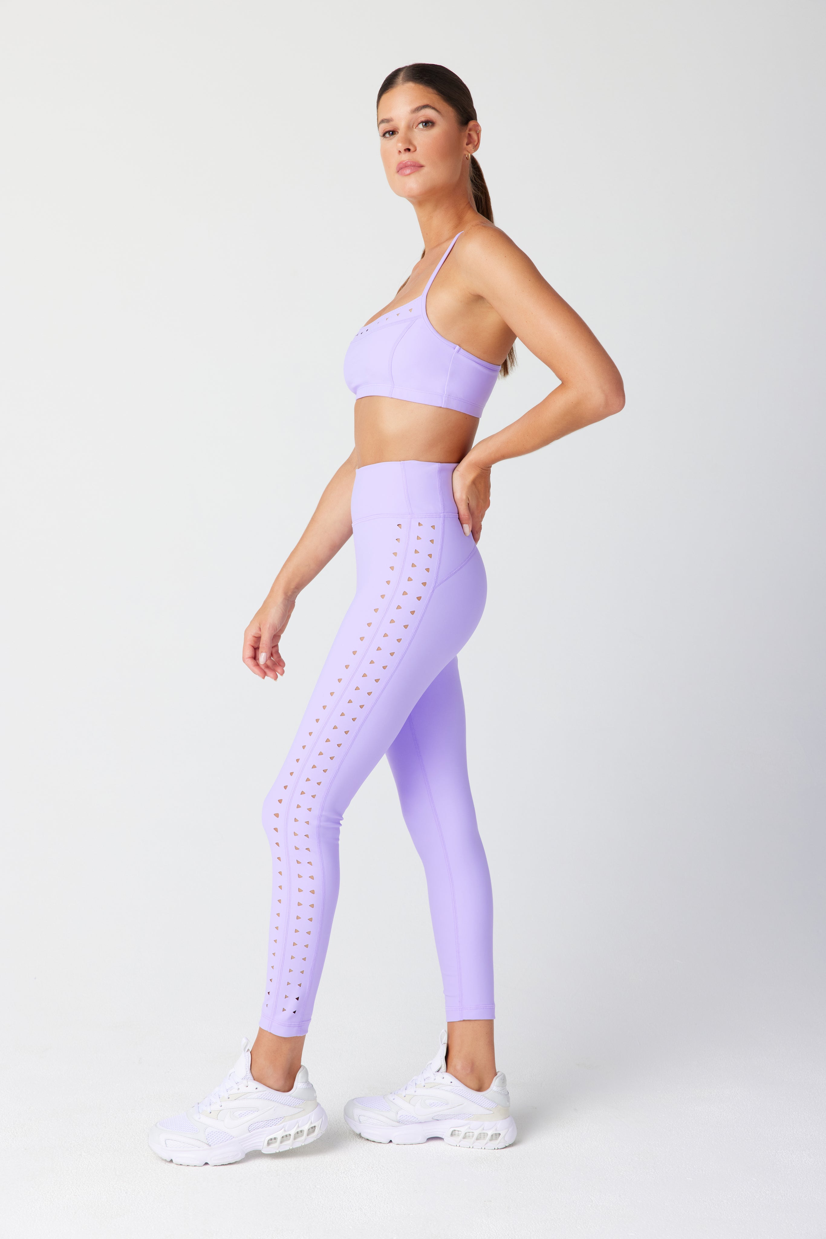 A person dressed in a lavender Gigi C Sport outfit, featuring the Giselle Bra with adjustable straps and recycled fabric leggings with cutouts, is wearing white sneakers and posing sideways against a plain white backdrop.