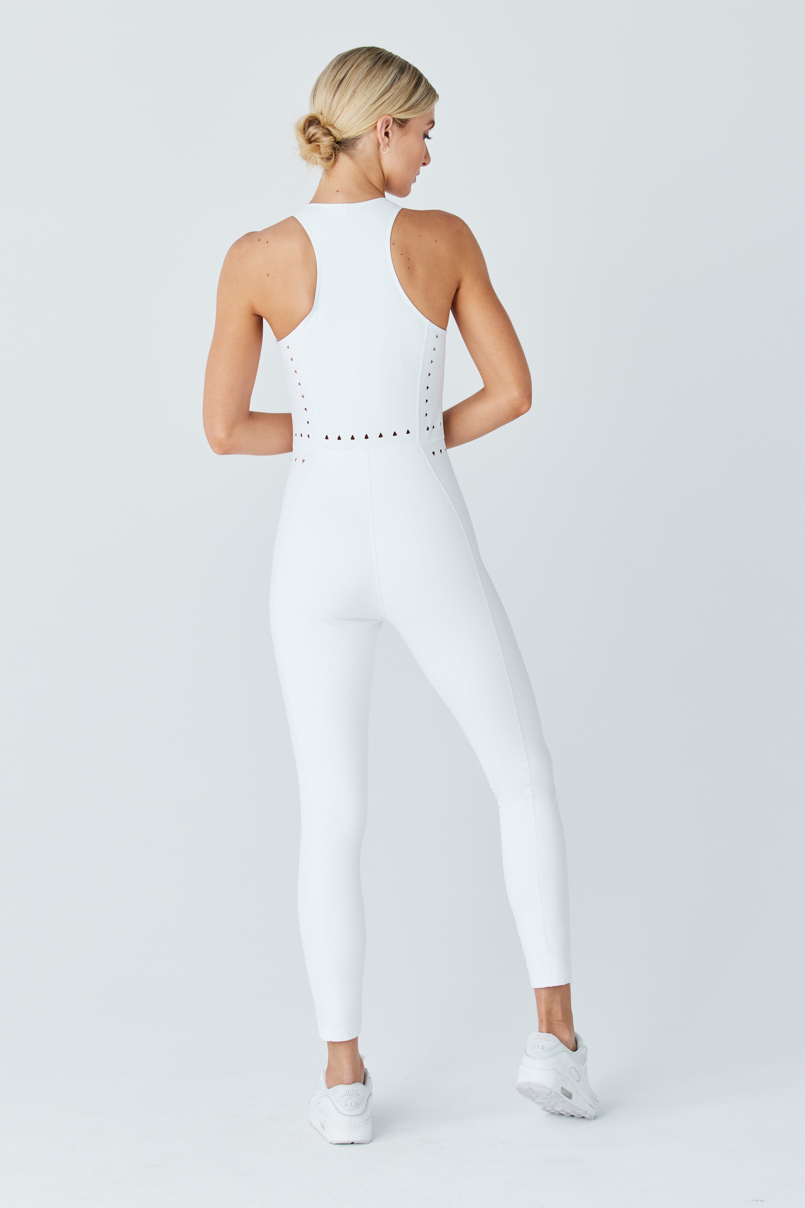 Wearing the Hunter Jumpsuit by Gigi C Sport, a person stands with their back to the camera in a white athletic jumpsuit and sneakers. The outfit features a racerback and cutout design, while they have styled their blonde hair in a bun against a plain background.