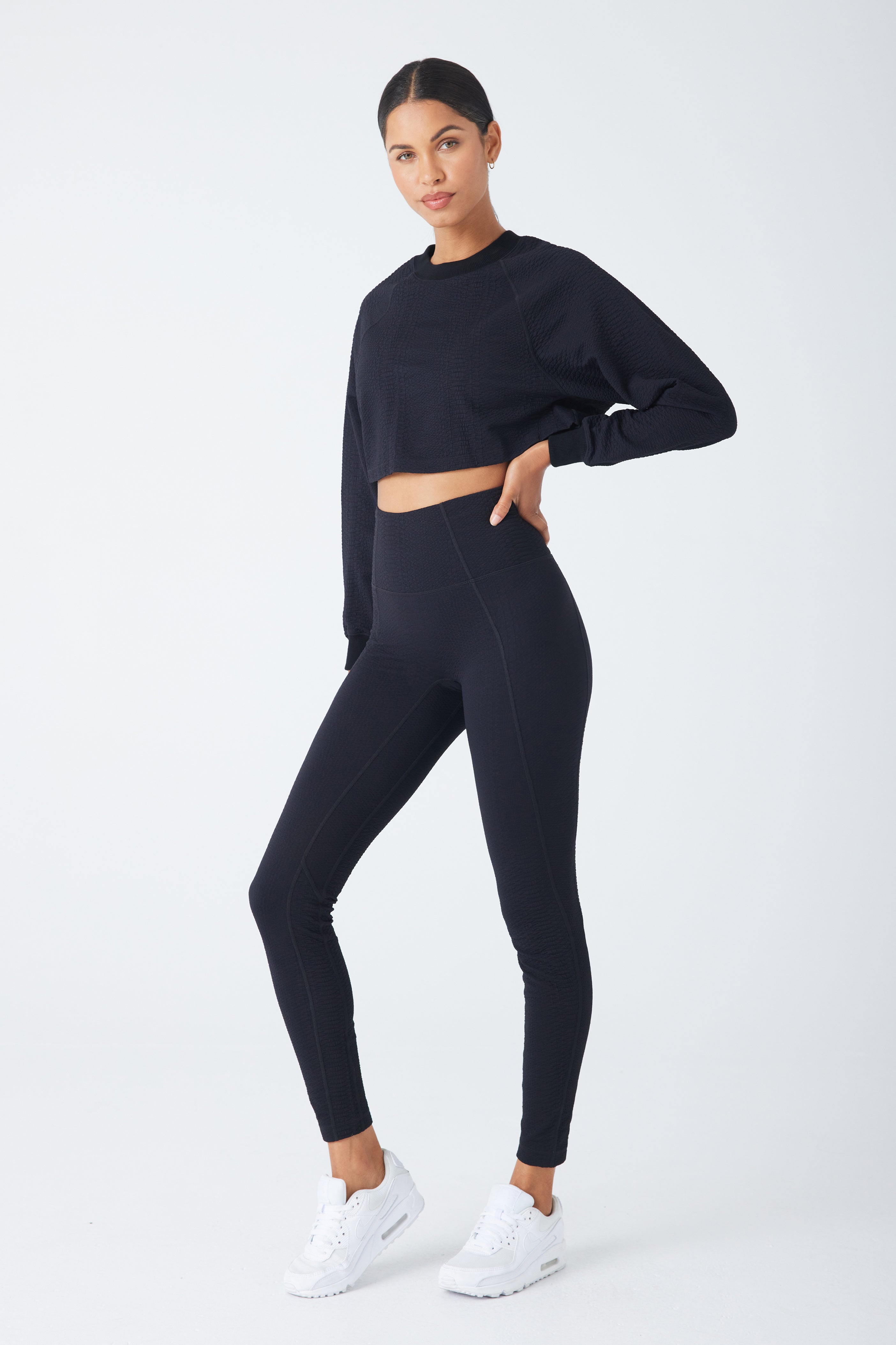 A person poses confidently in front of a white background, wearing a black cropped Isabella Pullover with a ribbed crew neck and matching leggings from Gigi C Sport, paired with white sneakers. One hand is on their hip while the other hangs by their side.