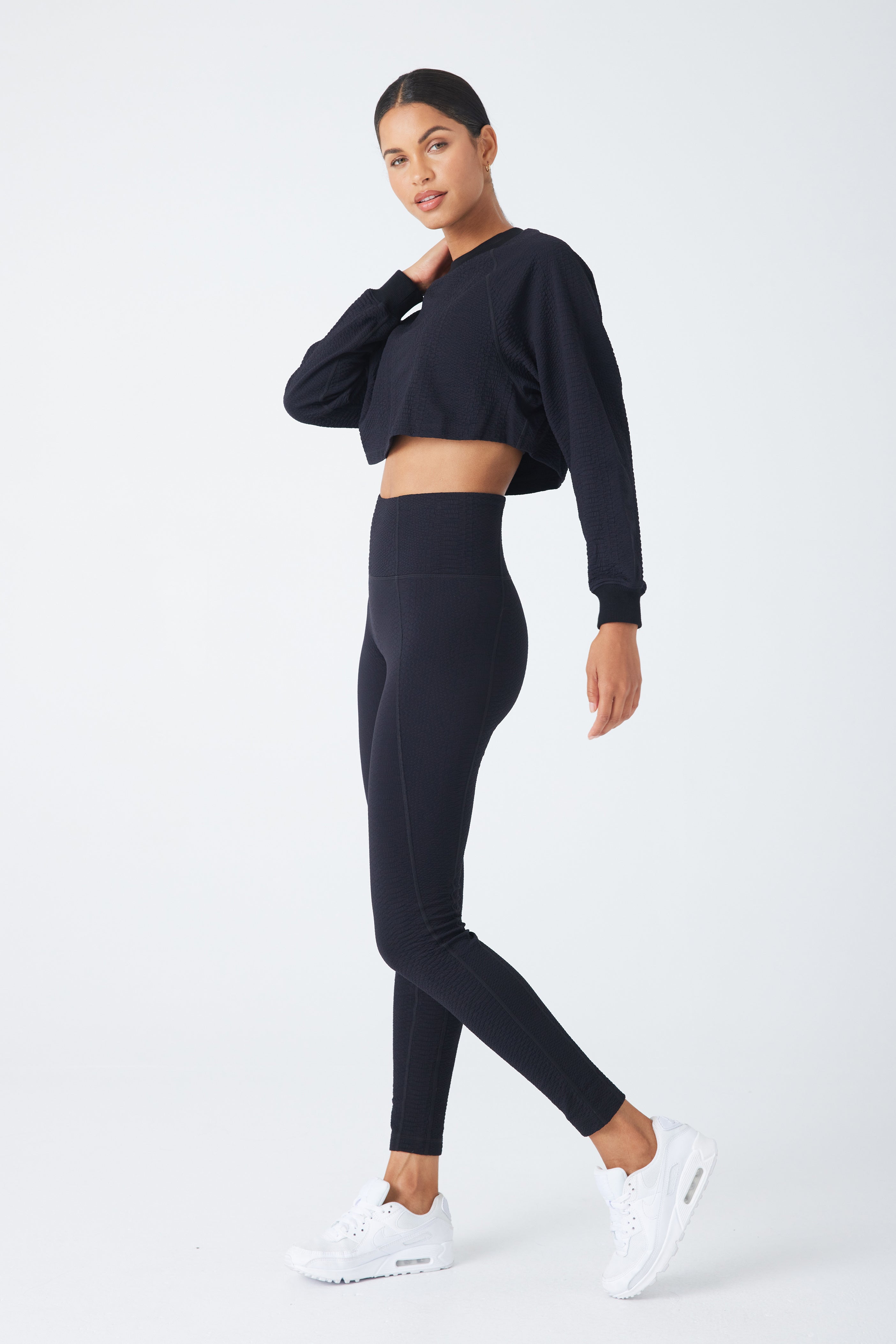 A woman confidently poses in the Isabella Pullover by Gigi C Sport, a black ribbed crew neck long-sleeve crop top, with high-waisted leggings and white sneakers. She stands on one leg, slightly turned to the side, against a plain white background.