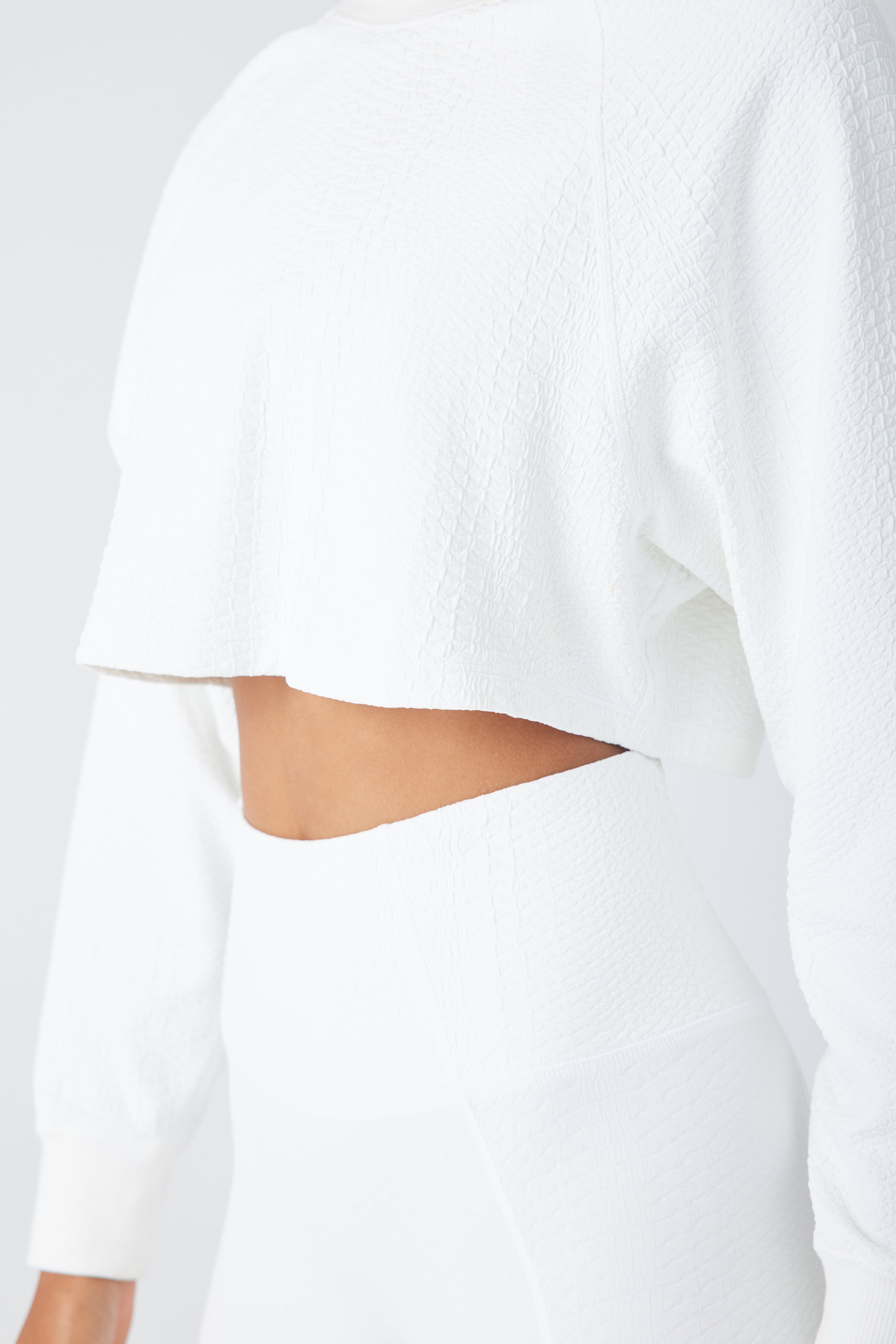 A person is wearing the Isabella Pullover by Gigi C Sport, a white textured long-sleeve cropped top with a ribbed crew neck, paired with high-waisted pants. The outfit looks soft and comfortable, emphasizing the midriff against a plain white background.