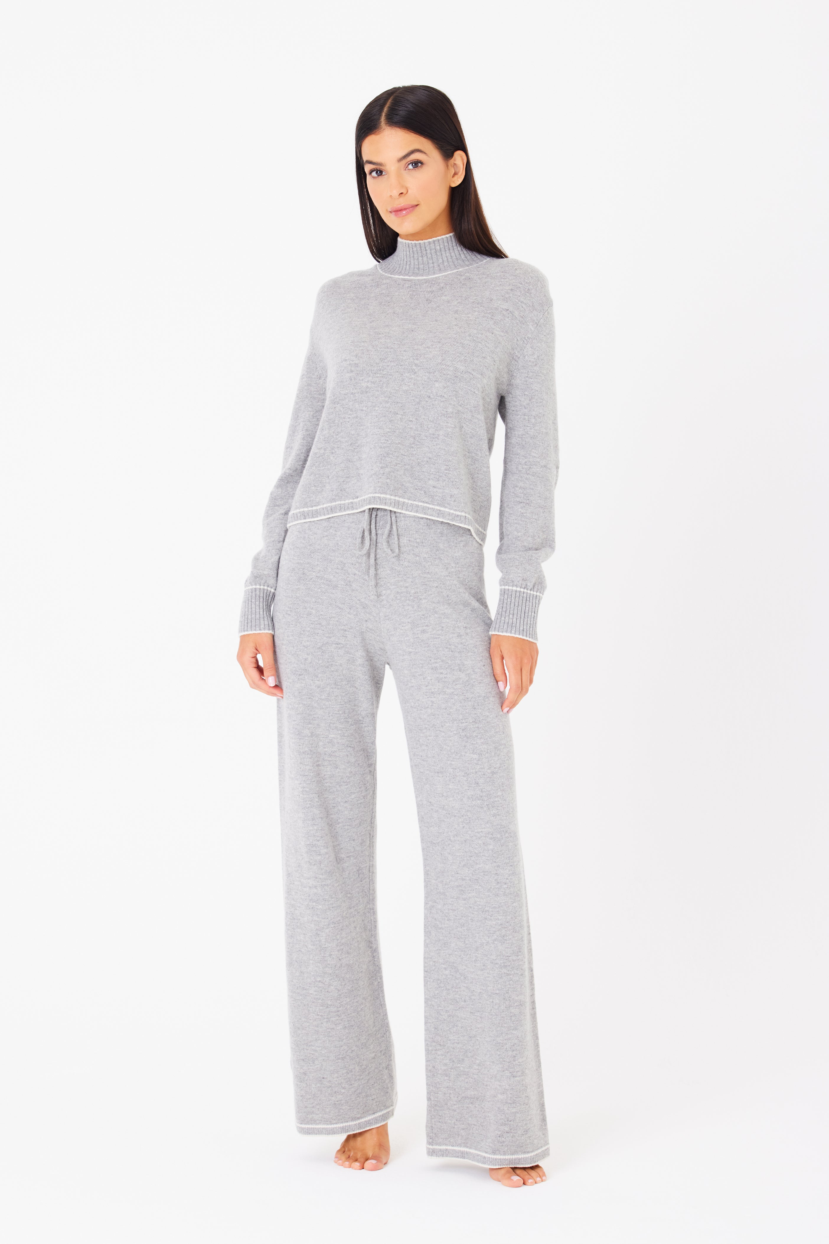 A woman in a Gigi C Lounge Jacqueline Pullover and wide-leg pants, made from recycled cashmere, stands barefoot against a white background. Her straight dark hair and neutral expression enhance the cozy feel of her outfit.