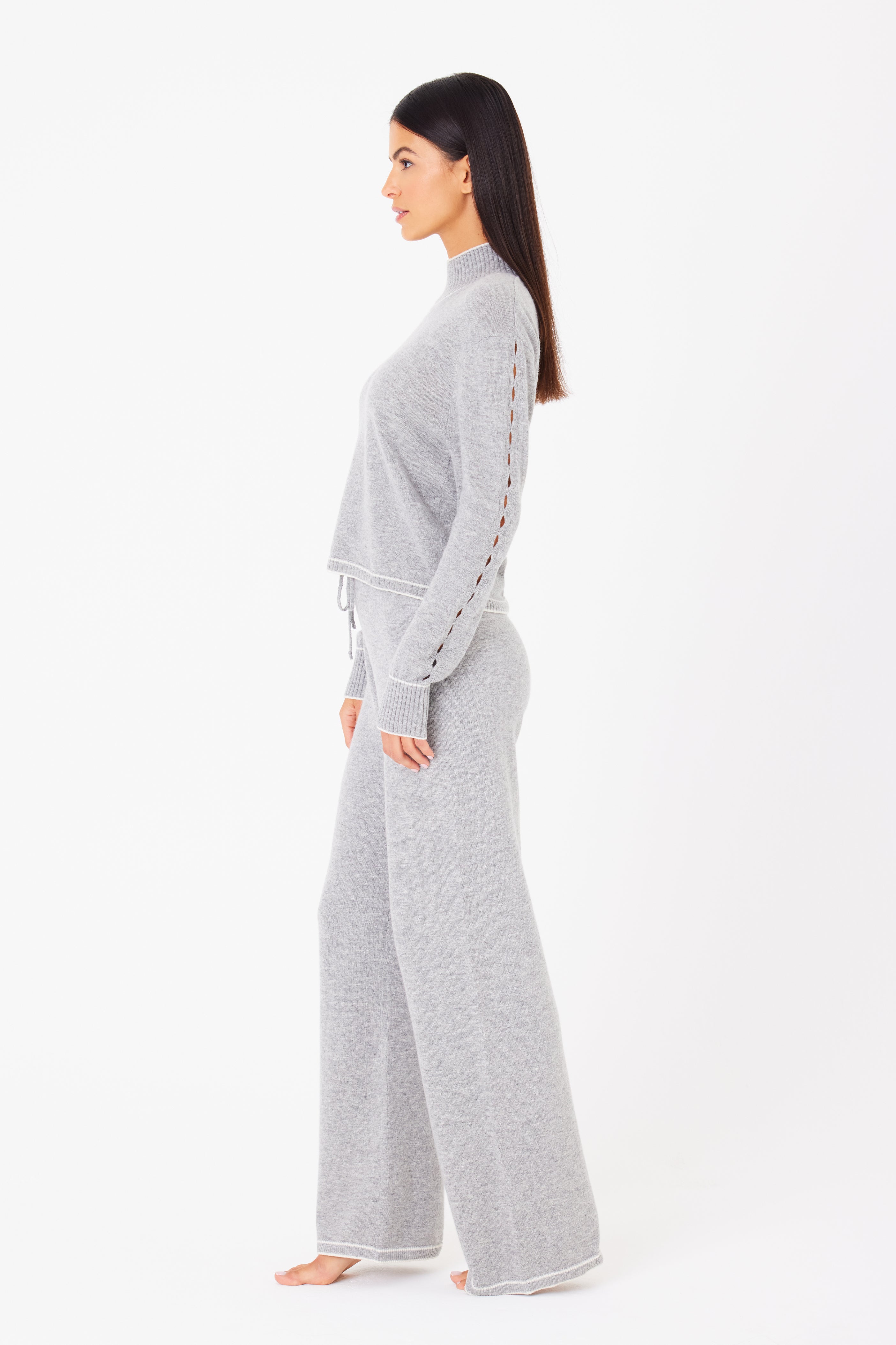 A person in a light gray wool cashmere knit sweater with decorative sleeve stitching and Aubrie Wide Leg Pant from Gigi C Lounge. The après-ski inspired outfit is barefoot against a white background, exuding effortless chic.