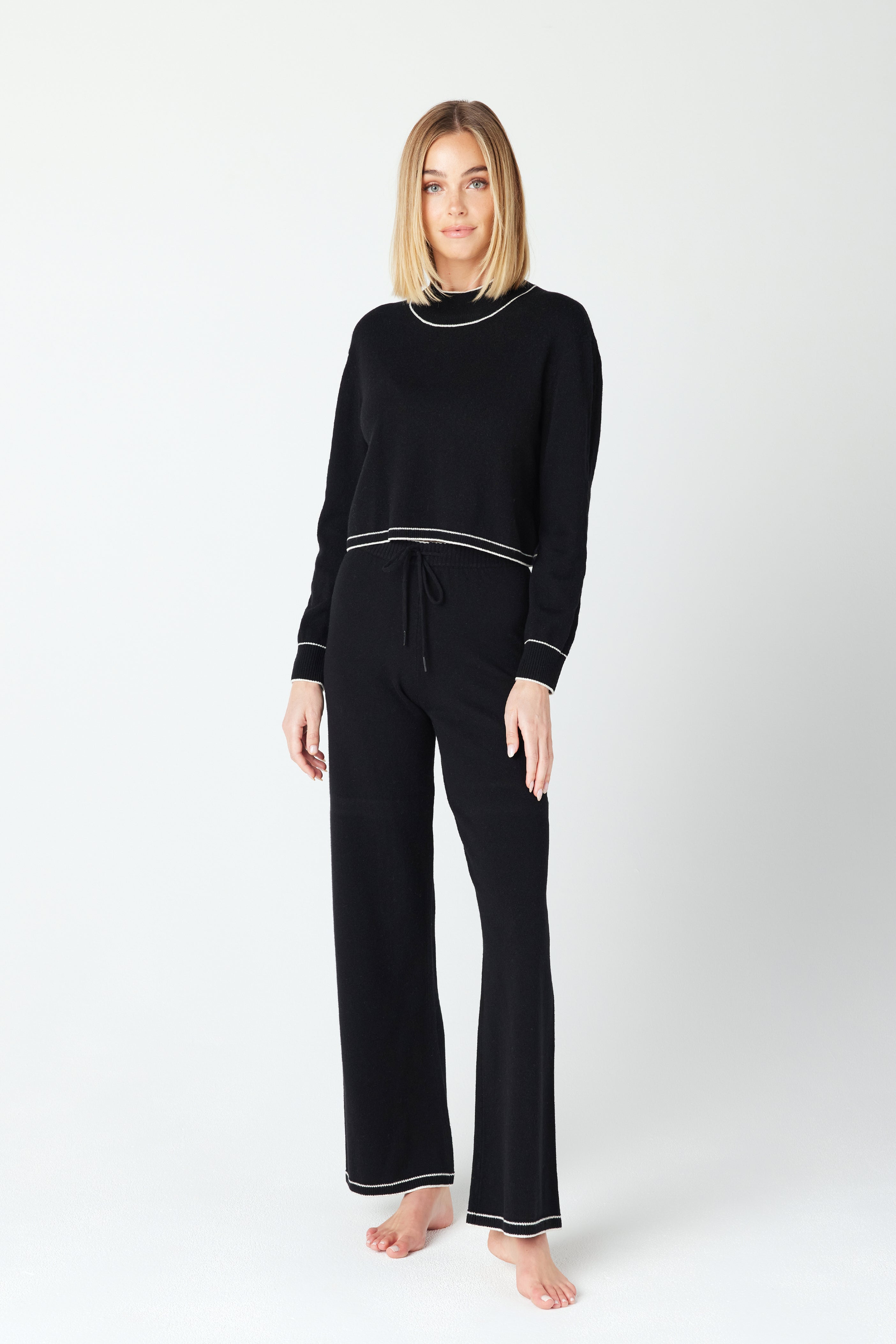 A person with shoulder-length hair models the Gigi C Lounge Jacqueline Pullover, a black recycled cashmere lounge set featuring matching wide-leg pants and white trim details, standing barefoot against a plain white background.