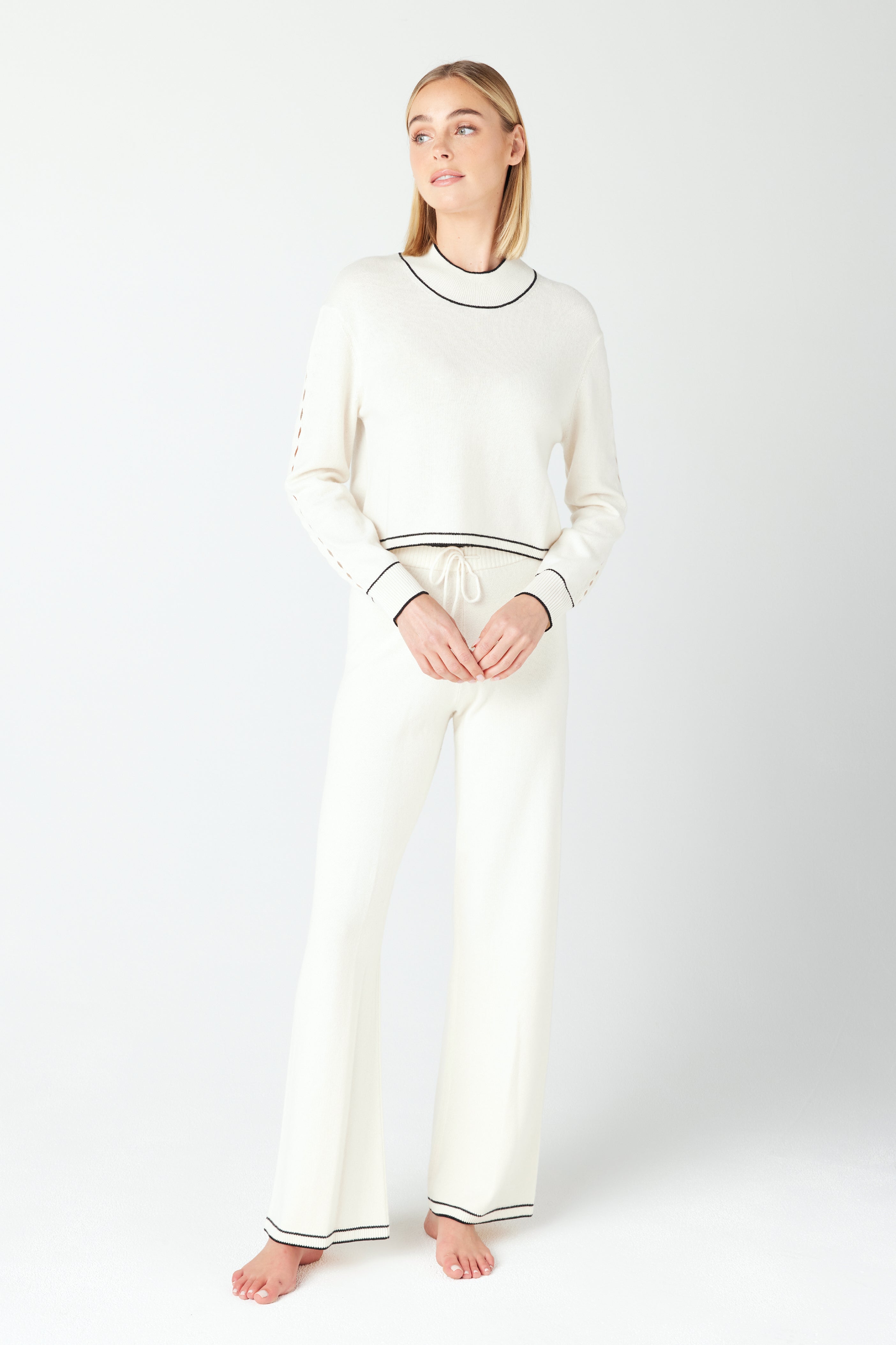 In a minimalist setting, someone stands barefoot in a matching white knit sweater and pants with black trim. Their Jacqueline Pullover by Gigi C Lounge showcases teardrop cut-out detailing, adding subtle elegance to the recycled cashmere look.